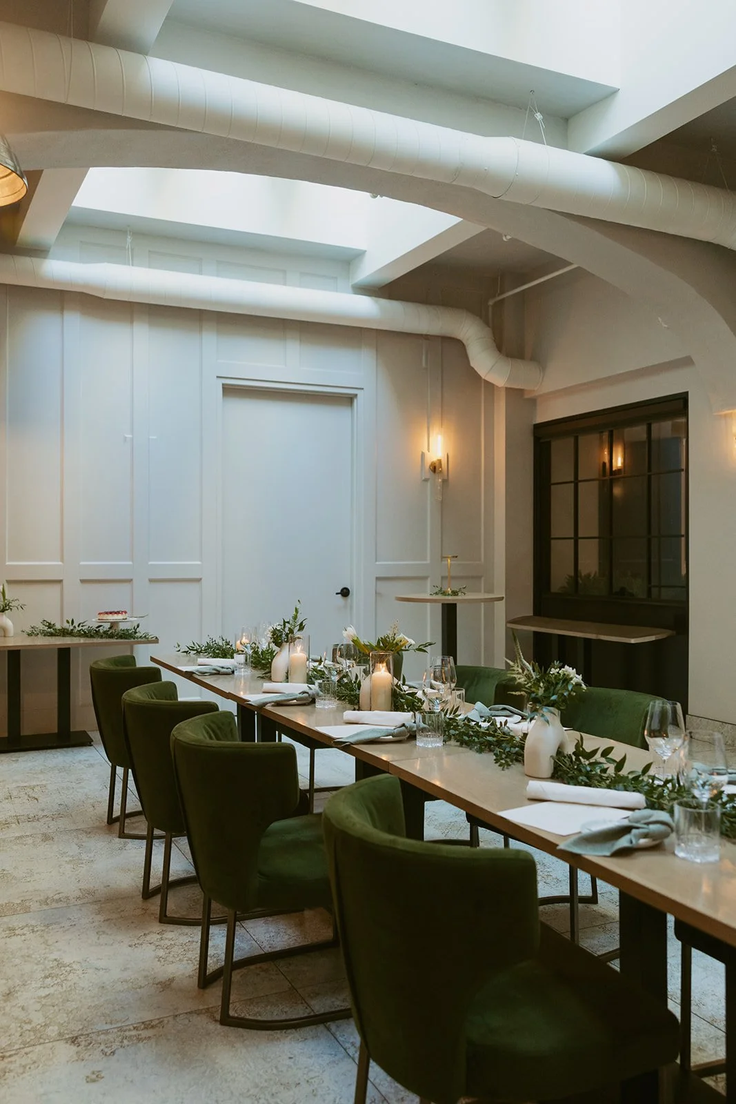 Interior of a modern dining room set up for a special event, with a long wooden table, green velvet chairs, candles, and floral centerpieces, in a well-lit space with white walls and ceiling.