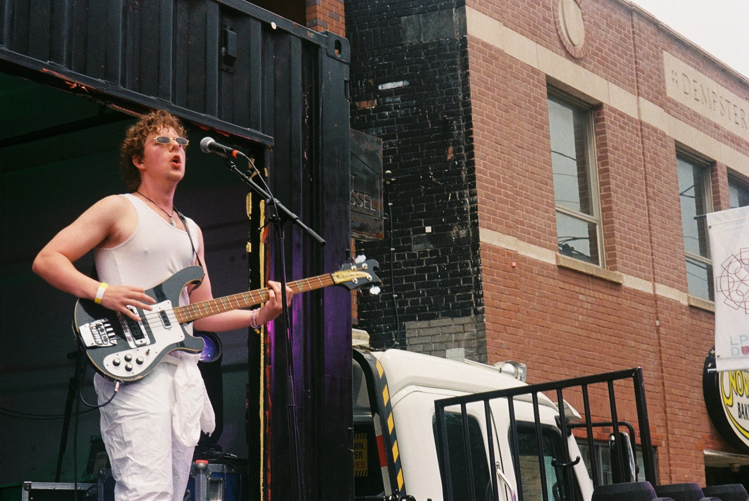 A musician performing on a stage outdoors, playing an electric guitar and singing into a microphone, with a brick building in the background.