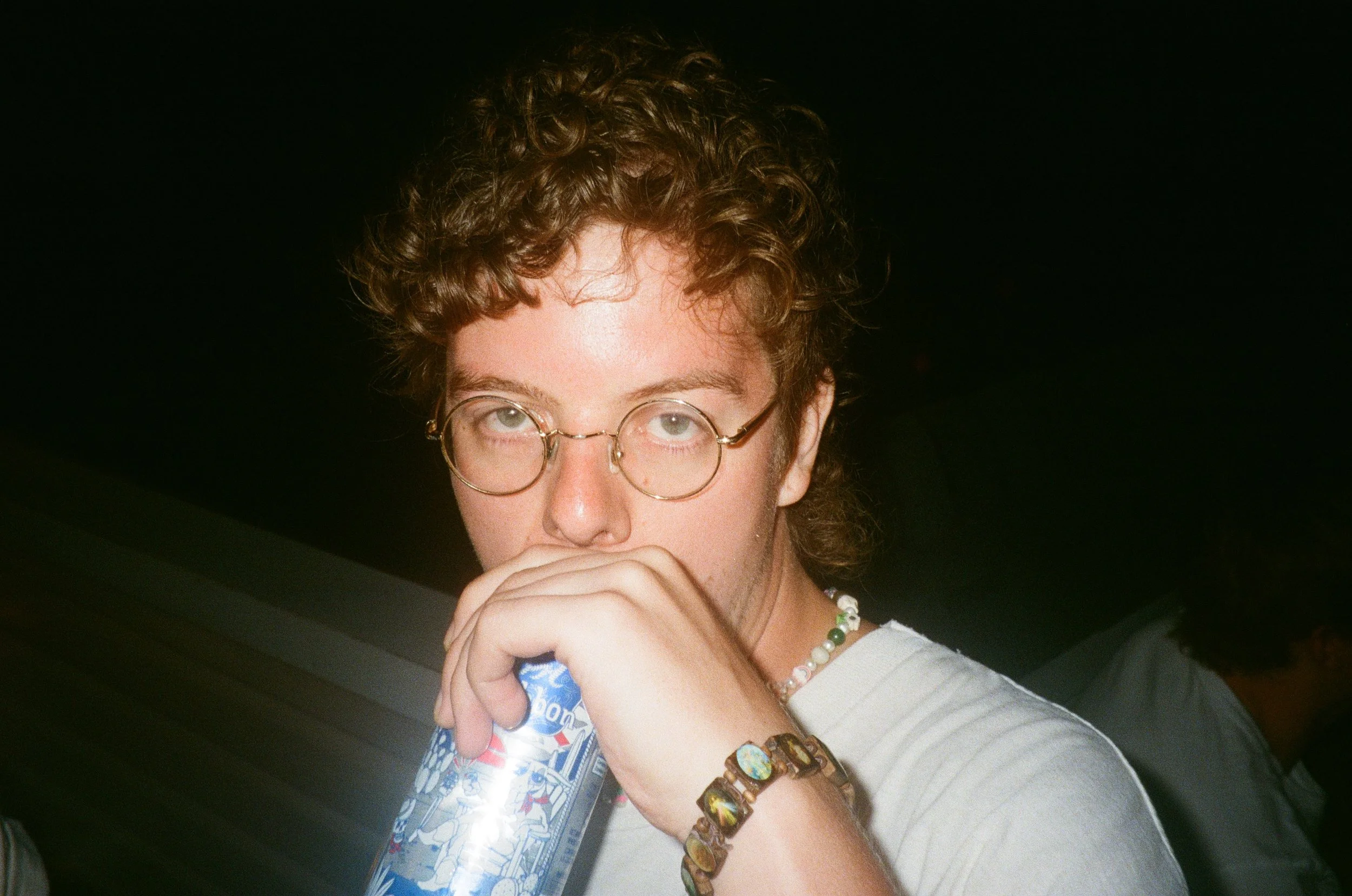 A young person with curly hair and glasses, holding a hand near their mouth, wearing necklaces and bracelets, against a dark background.