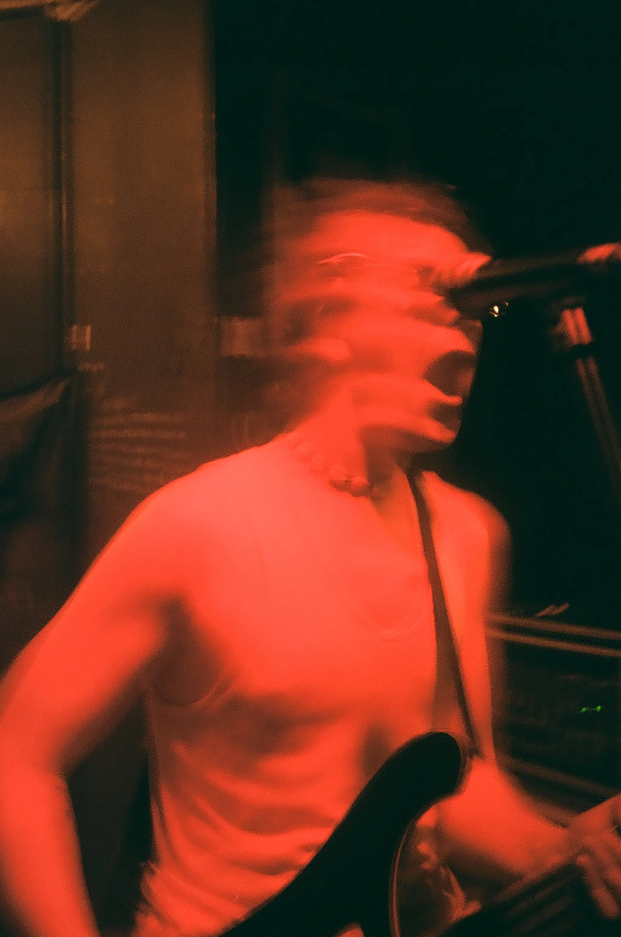 A person with long hair wearing an orange shirt, possibly singing or speaking into a microphone, in a dark environment with red lighting.