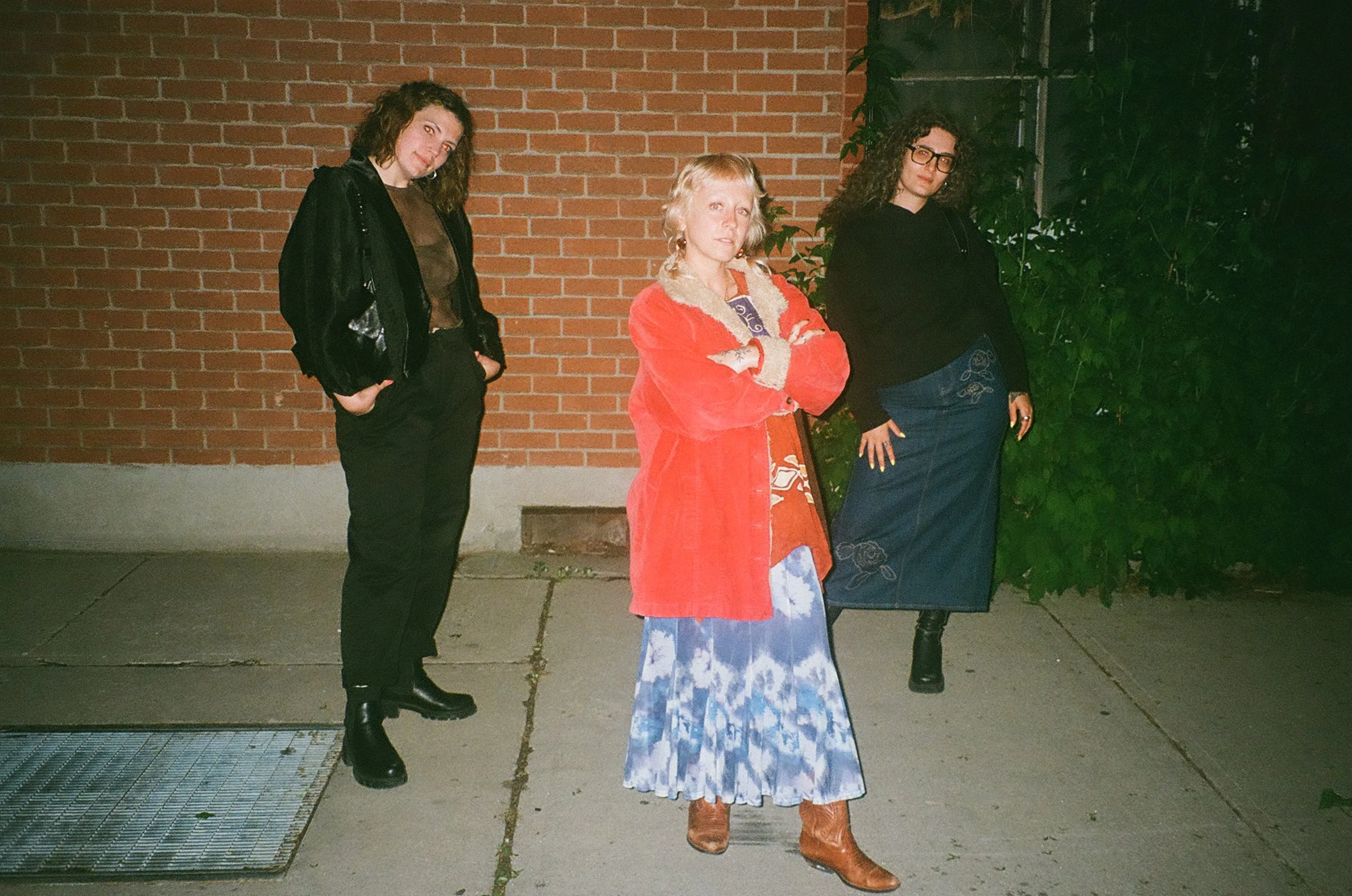 Three women standing outside against a brick wall and greenery at night. The woman on the left has curly hair, wearing a black jacket and black pants. The woman in the middle has short blonde hair, wearing a red jacket and a dress with a blue and white pattern. The woman on the right has curly hair, glasses, a black jacket, and a long blue skirt with embroidery.