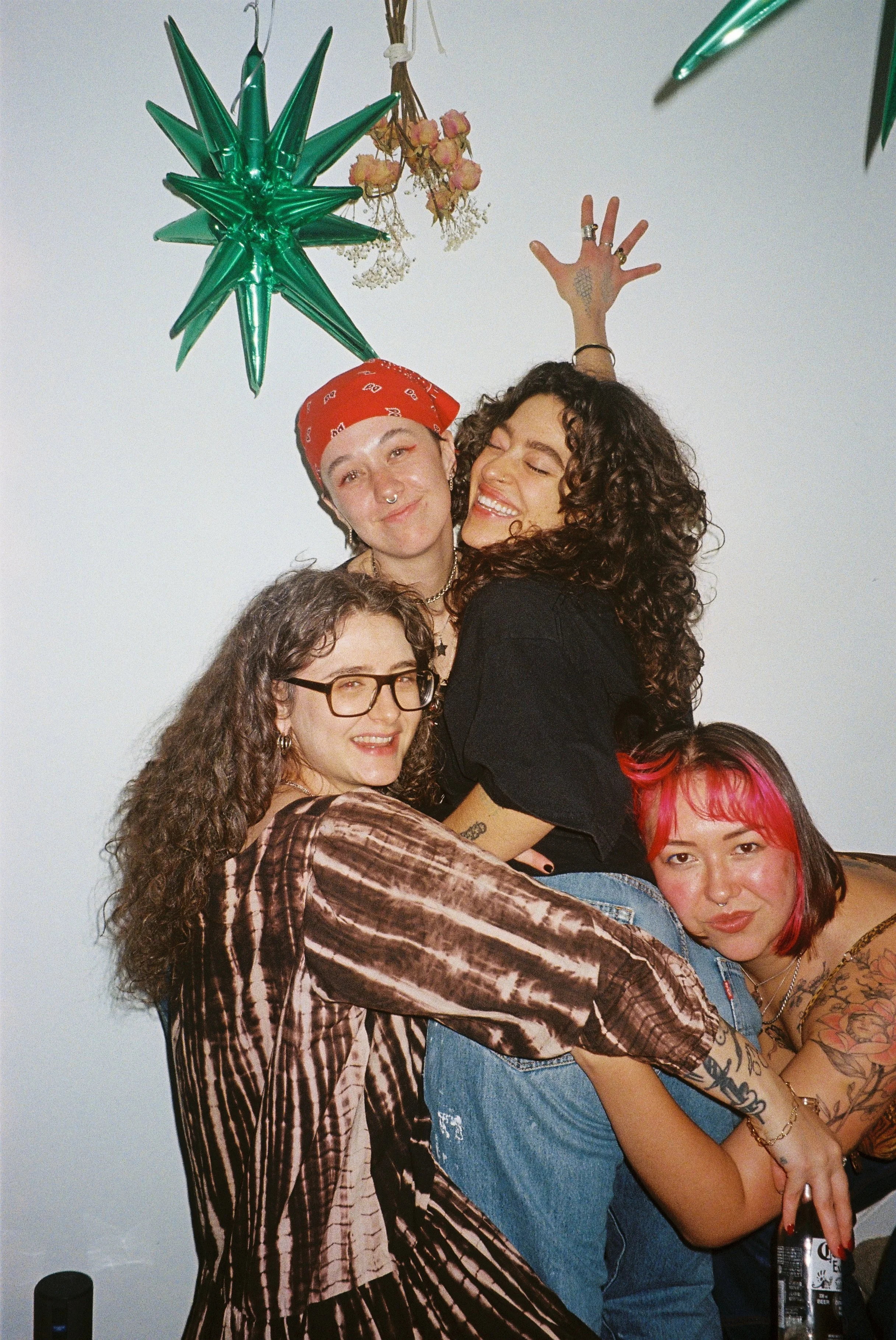 Group of five friends celebrating together, with some wearing colorful clothing, including a zebra print dress, denim, and tattoos visible. They are smiling and hugging, with a white wall background and festive decorations like a green star-shaped balloon and dried flowers hanging from the ceiling.