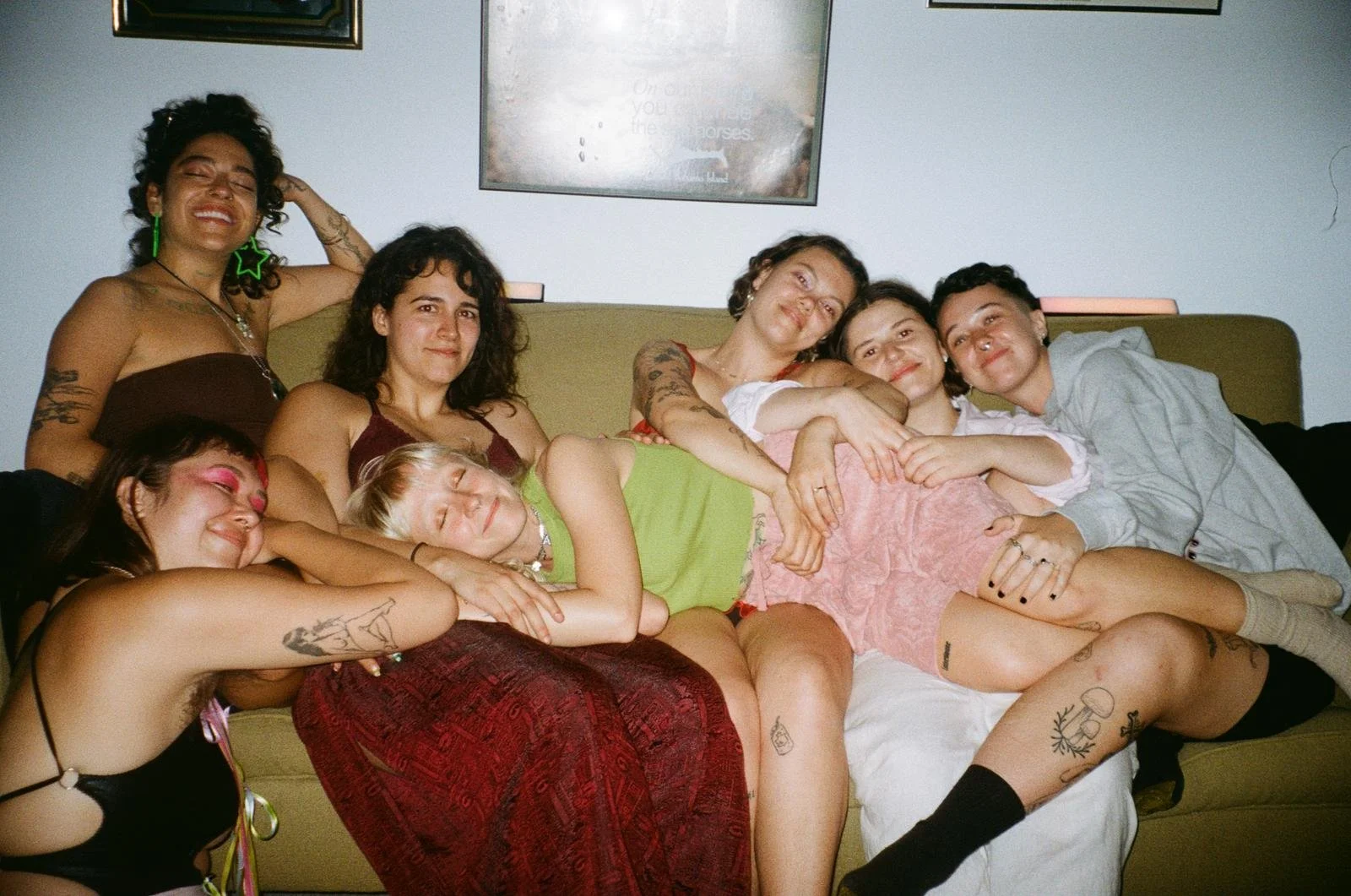 A group of eight women sitting and lying on a green couch, smiling and hugging, in a cozy indoor setting with framed pictures on the wall behind them.