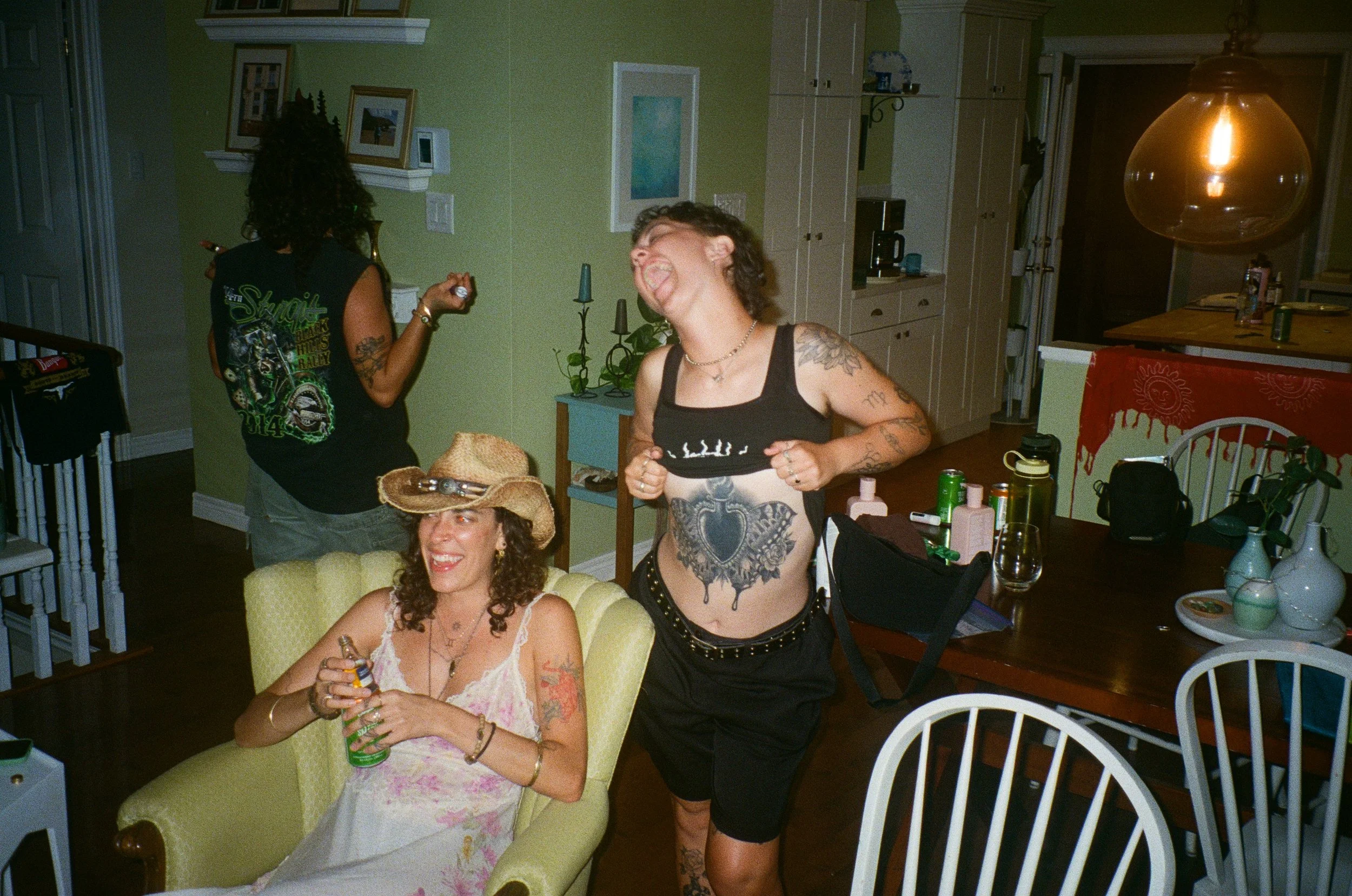 Two women are in a kitchen and living room area. One woman is sitting on a yellow chair, holding a can and smiling. The other woman stands, appears to be mid-laugh, and has tattoos on her arms and torso. There are various objects and decorations around them, including a large hanging light, framed pictures, and potted plants.