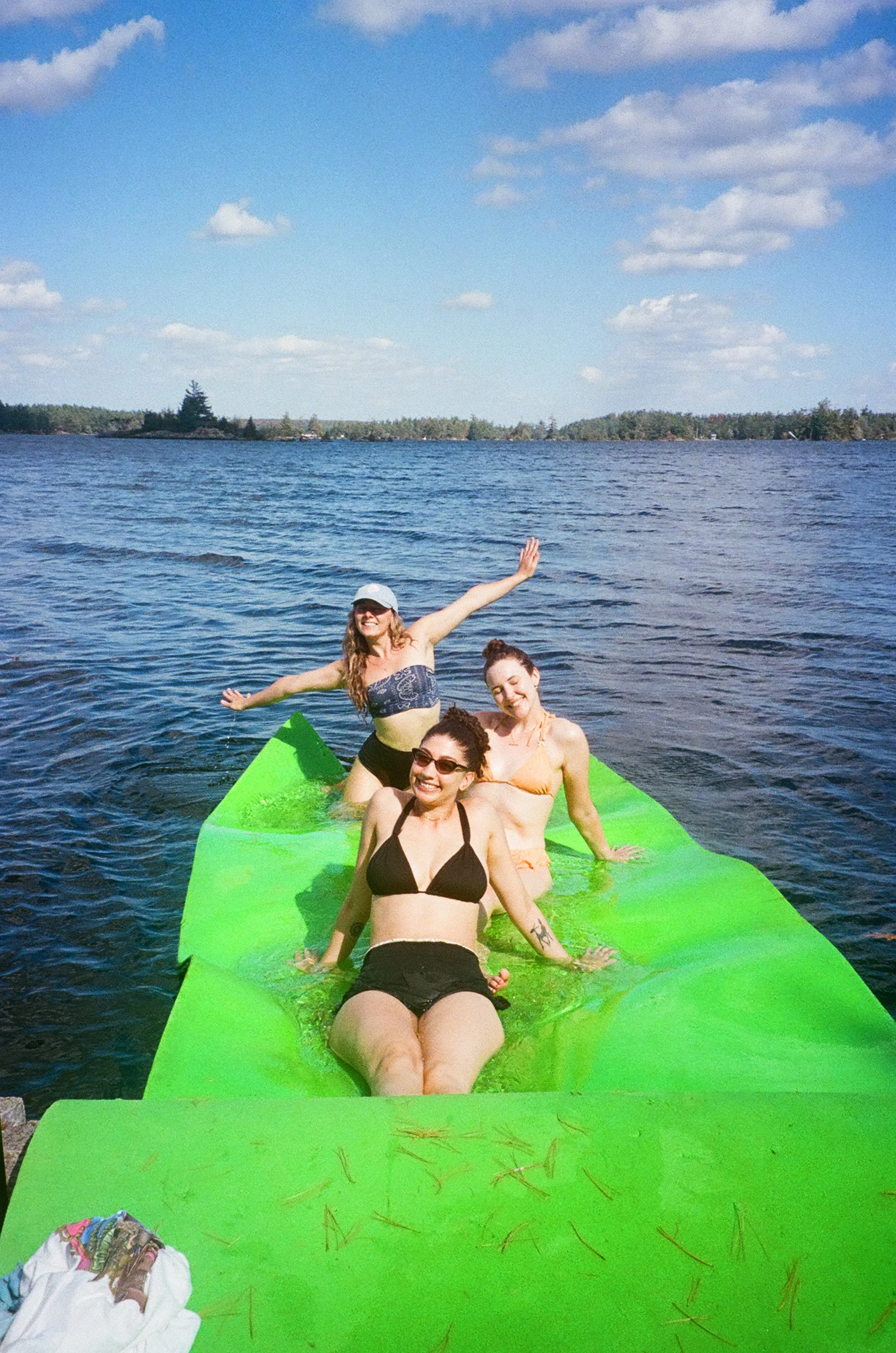 Four women are sitting on a large green floating object in a lake, enjoying sunny weather. They are smiling and appear to be having fun, wearing swimsuits and casual beach clothing. The background features a lake with treetops and a partly cloudy sky.