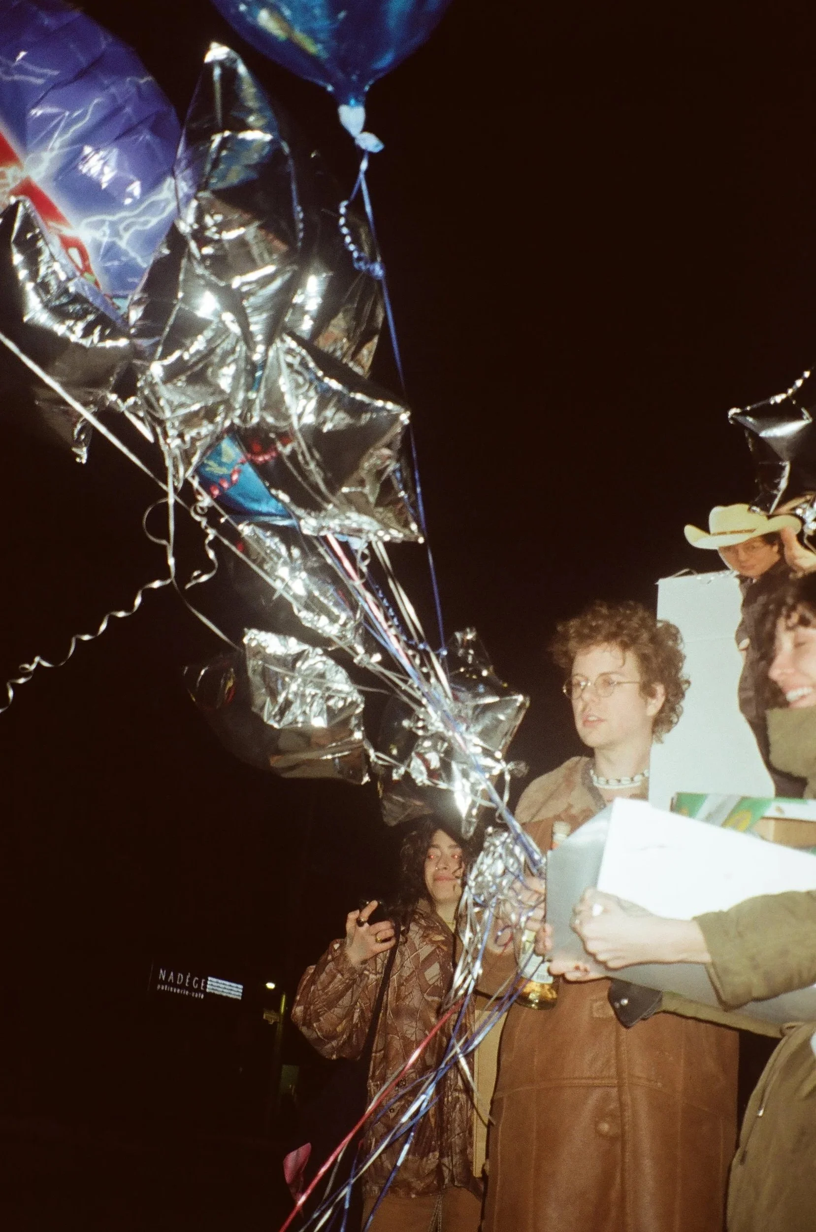 Group of people at a night event, surrounded by metallic balloons and colorful ribbons, some holding papers and wearing casual clothing and cowboy hats.