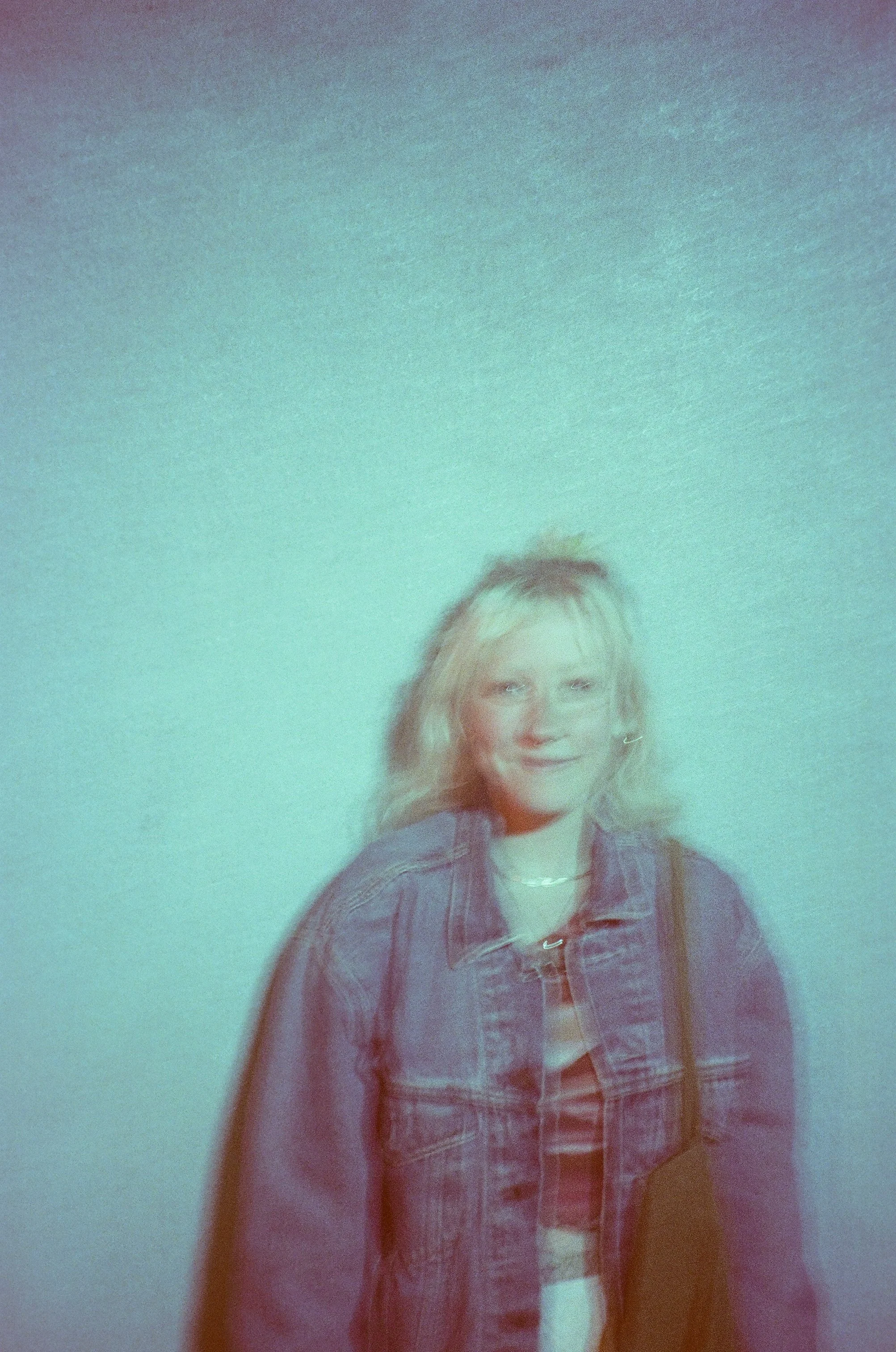 A young woman with long, light-colored hair smiling, wearing a denim jacket over a plaid shirt, standing against a light green wall.