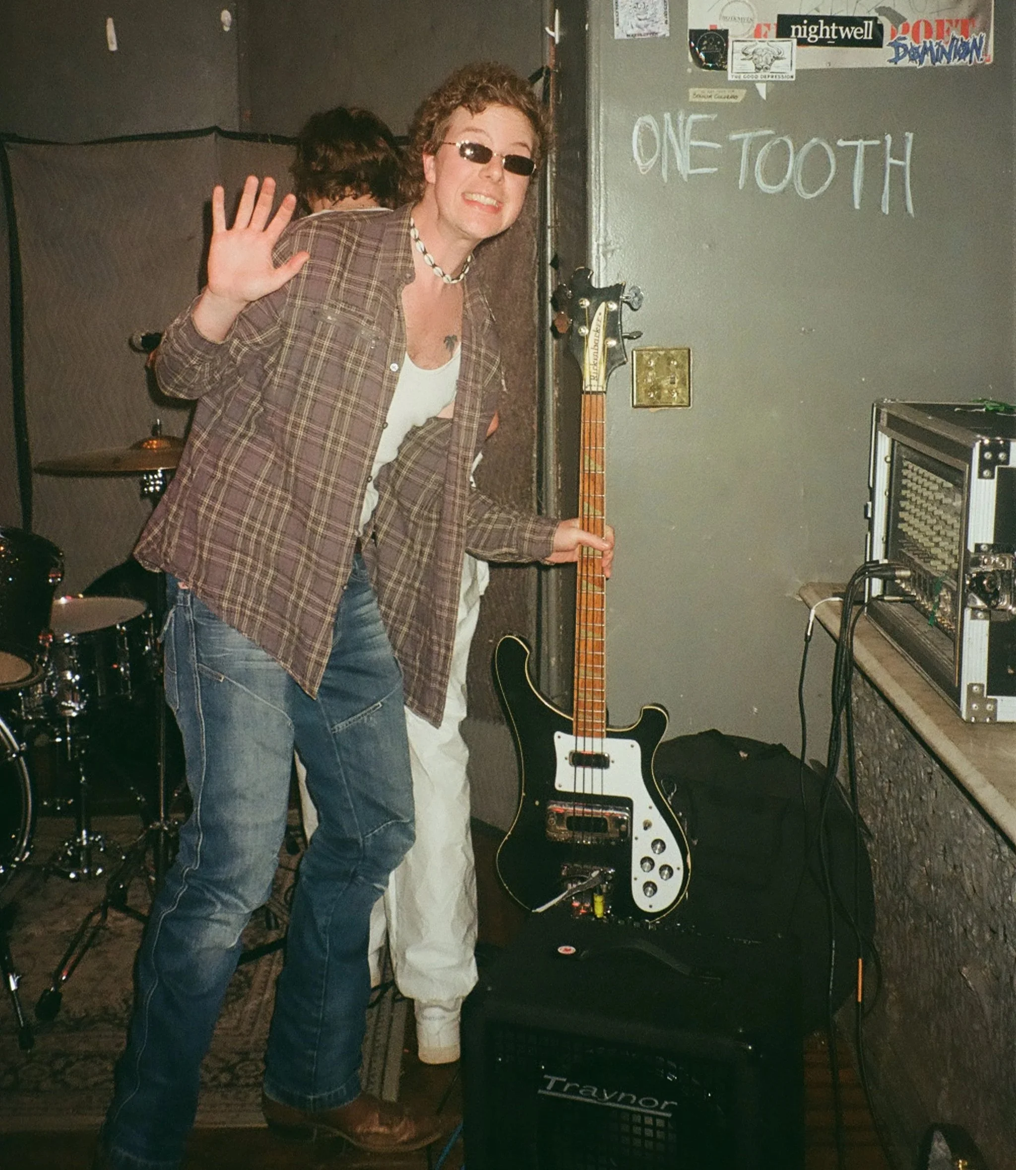 Person waving in a music rehearsal space, holding a black and white electric guitar, with musical equipment and band posters on the wall.