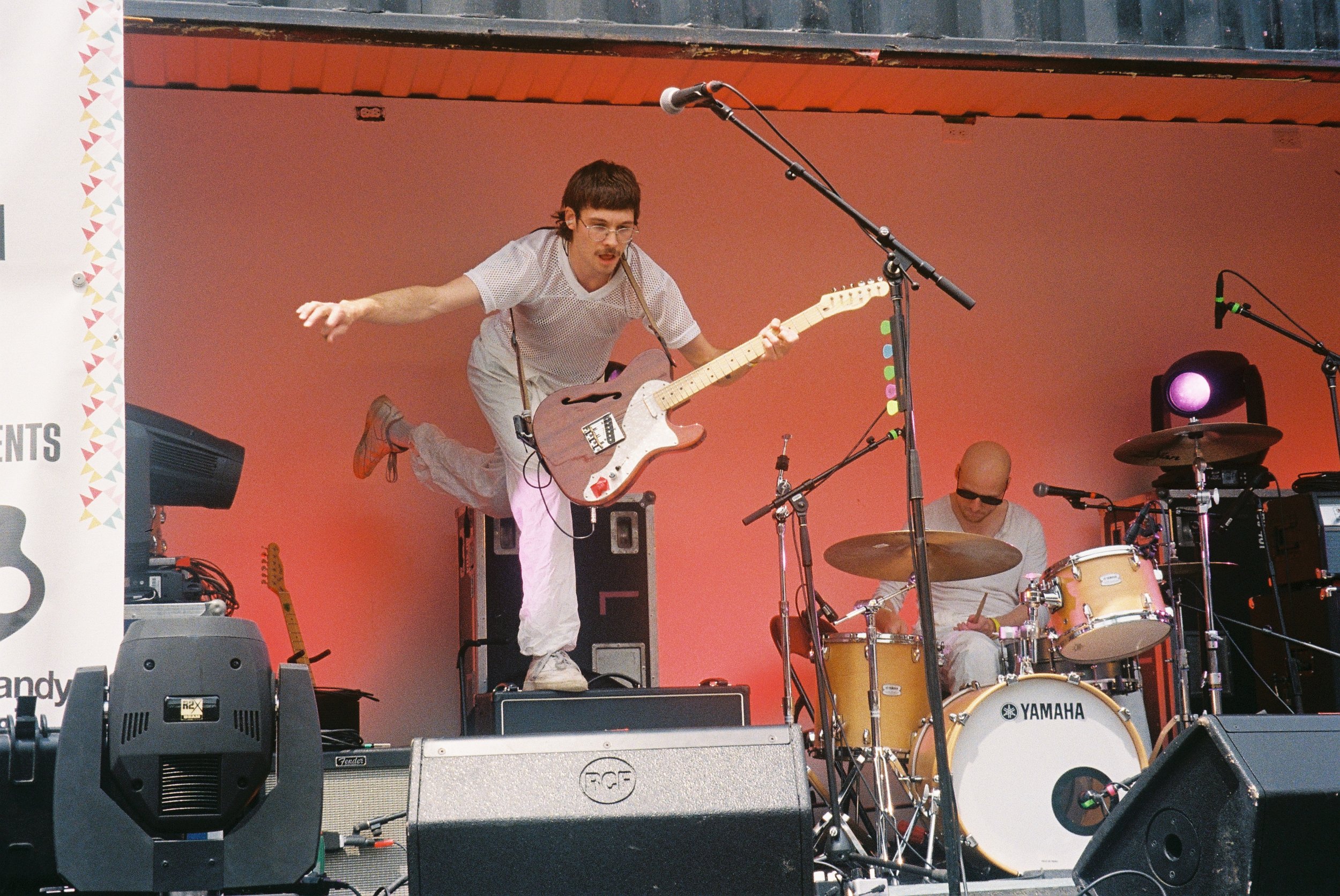 A musician jumping with a guitar on stage during a performance, with other band members playing drums and bass in the background. The stage has a pinkish background and various audio equipment and microphones are visible.