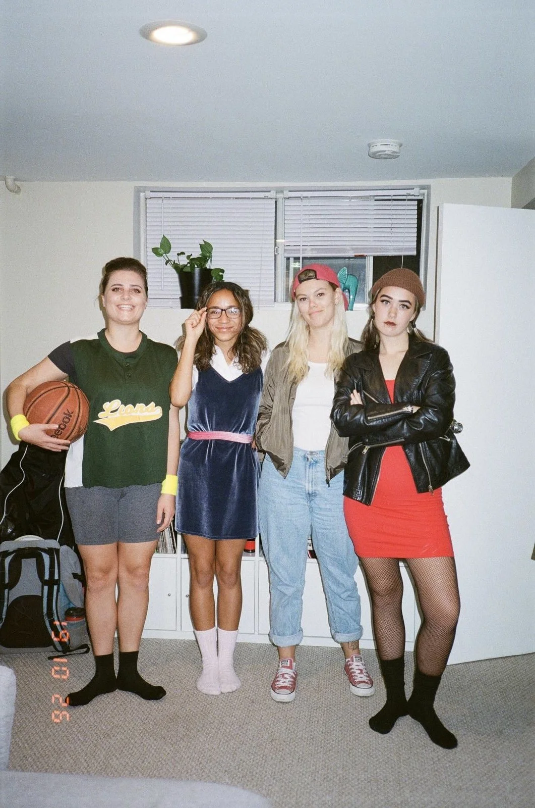 Four young women standing side by side indoors, one holding a basketball, dressed in casual and sporty outfits with a mix of serious and cheerful expressions.