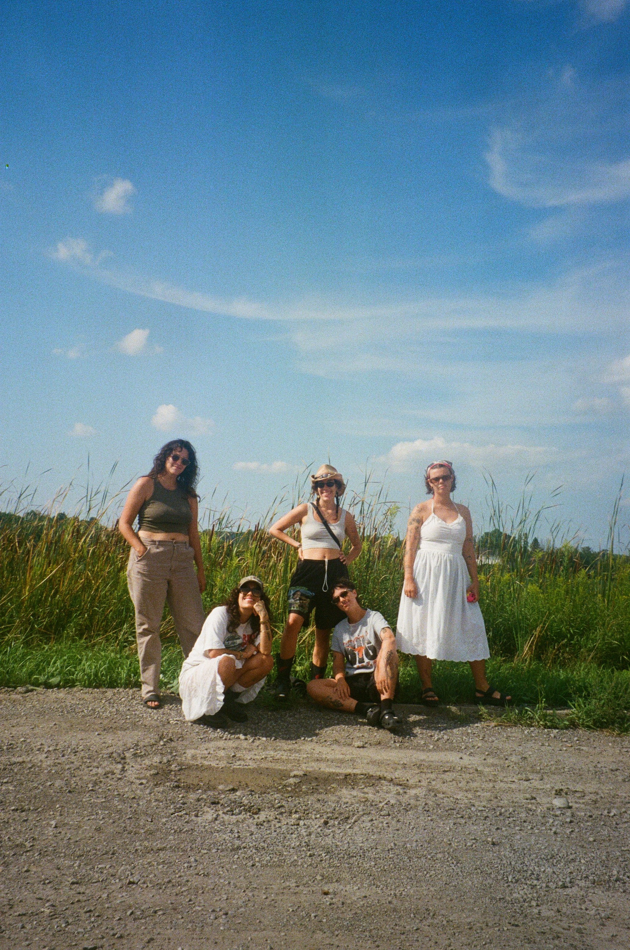 A group of seven young people outdoors on a sunny day with a blue sky and scattered clouds, standing and sitting on a patch of grass and dirt, some wearing sunglasses and casual summer clothing.