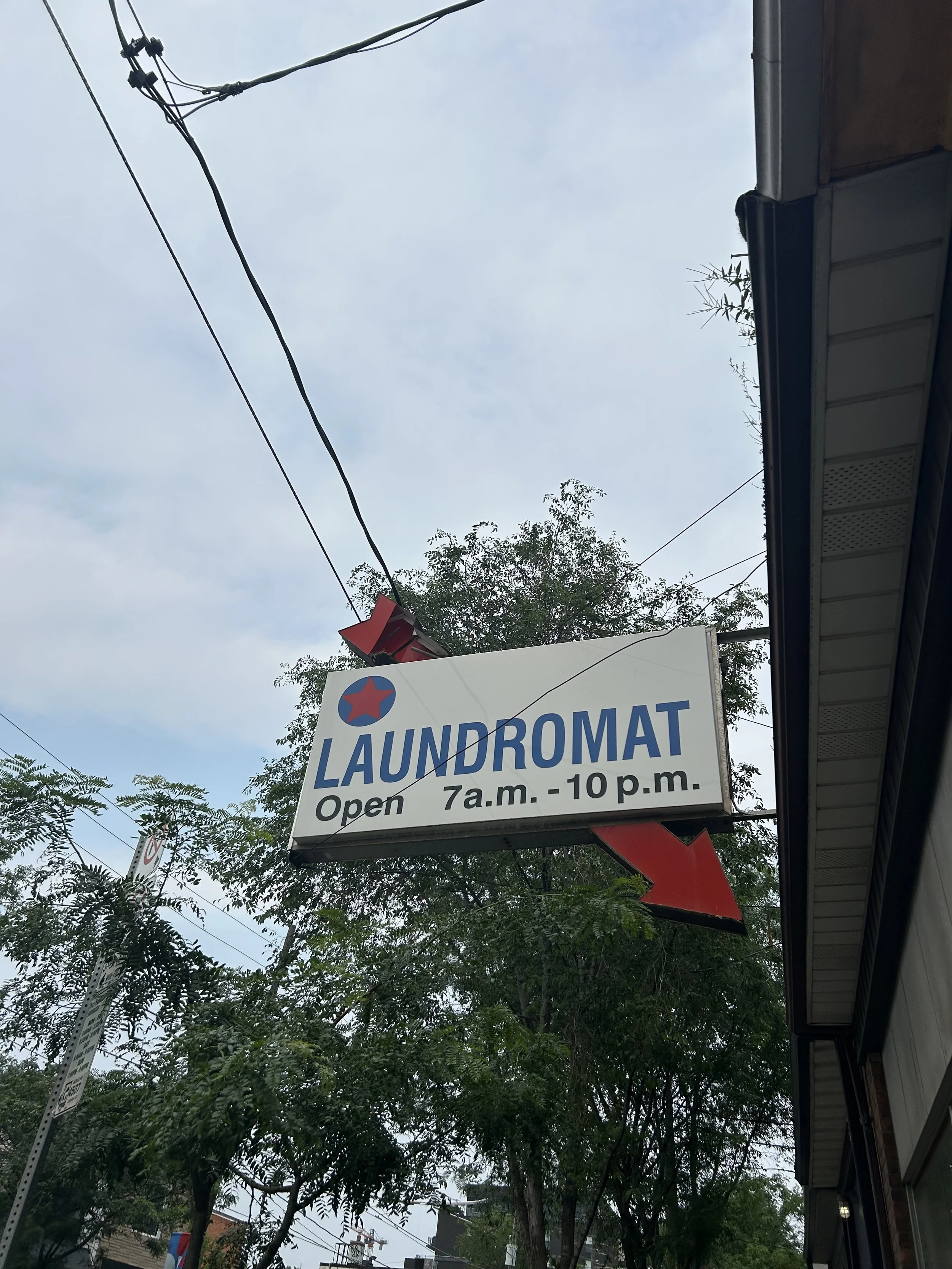 A street view showing a sign for a laundromat open from 7 a.m. to 10 p.m., with trees and power lines in the background, and part of a building roof on the right side of the image.