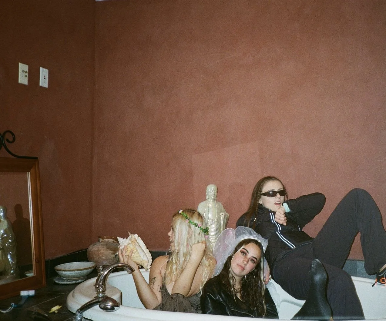 Three young women sitting and lying in a bathroom bathtub, with a pinkish wall, a mirror, and decorative items like a statue and patterned items around them.