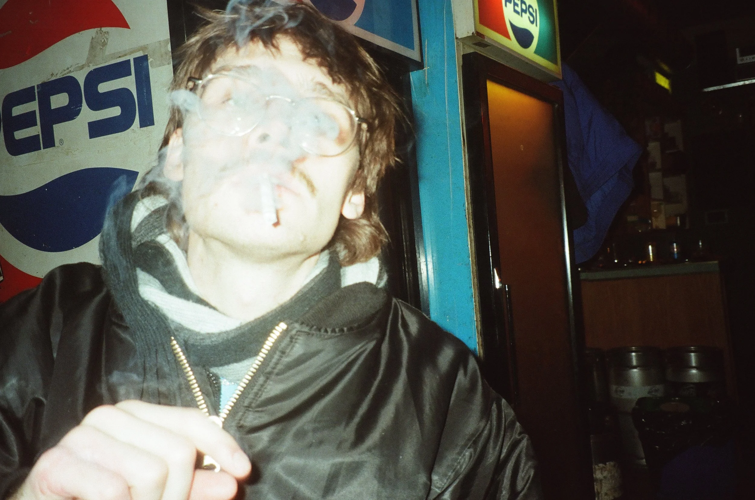 A young man with glasses and curly brown hair, smoking a cigarette, wearing a black jacket and striped sweater, standing in front of a vending machine with Pepsi branding at night.
