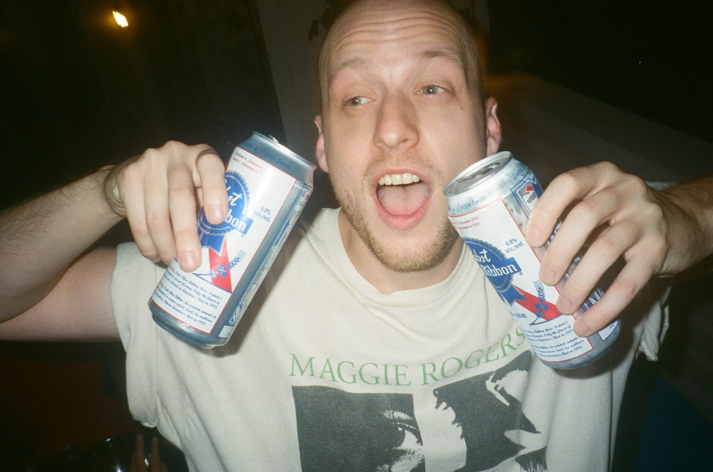 A man holding two cans of Pabst Blue Ribbon beer, one in each hand, with his mouth open as if shouting or singing, wearing a gray T-shirt with a graphic of Maggie Rogers.