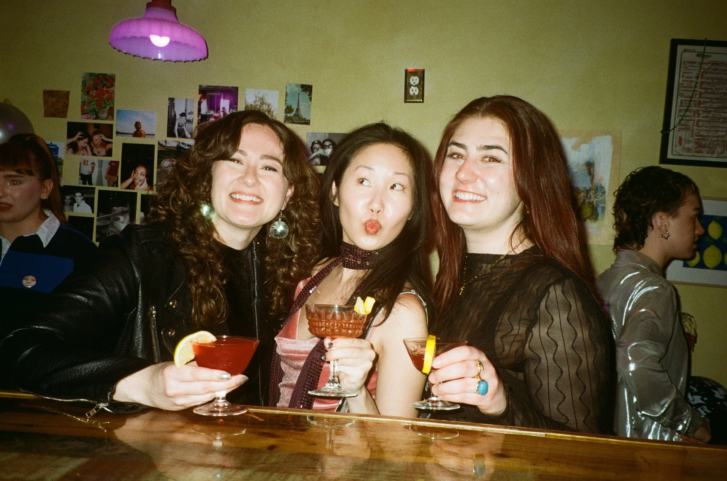 Three women smiling and holding drinks at a social gathering. The woman on the left has curly hair and is wearing a black jacket and large earrings. The middle woman with straight hair is making a kissy face, and the woman on the right has red hair and is smiling. Behind them is a wall with photos and artwork, a purple hanging lamp, and people to the right side of the image.