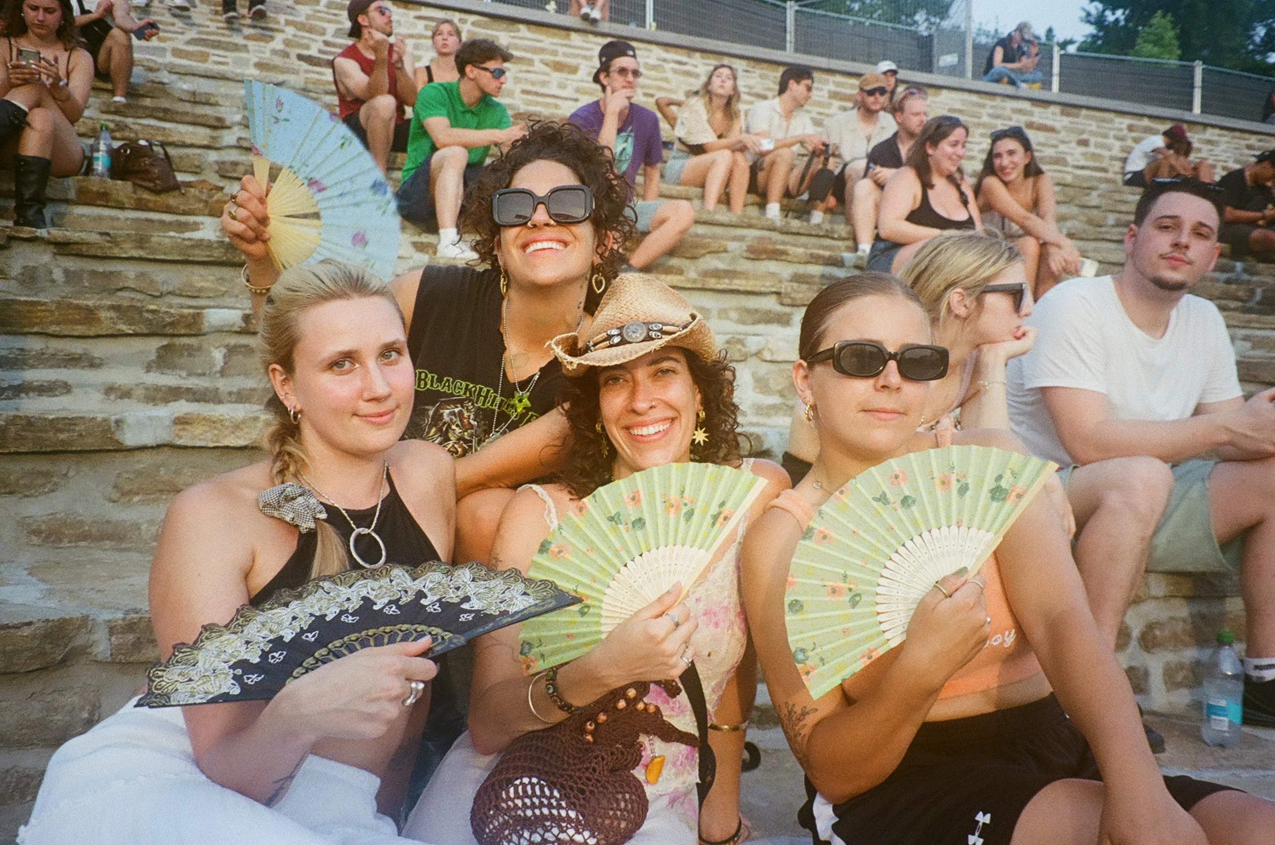 A group of people sitting on stone steps outside, some holding colorful fans, with a woman in the center wearing sunglasses and smiling.