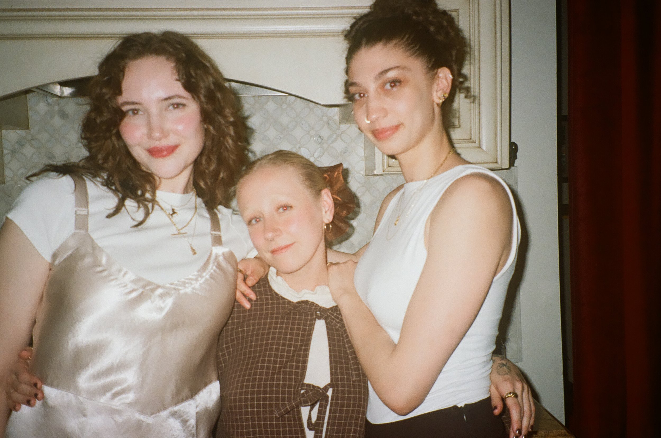 Three women with curly and straight hair posing closely together in an indoor setting, smiling at the camera.