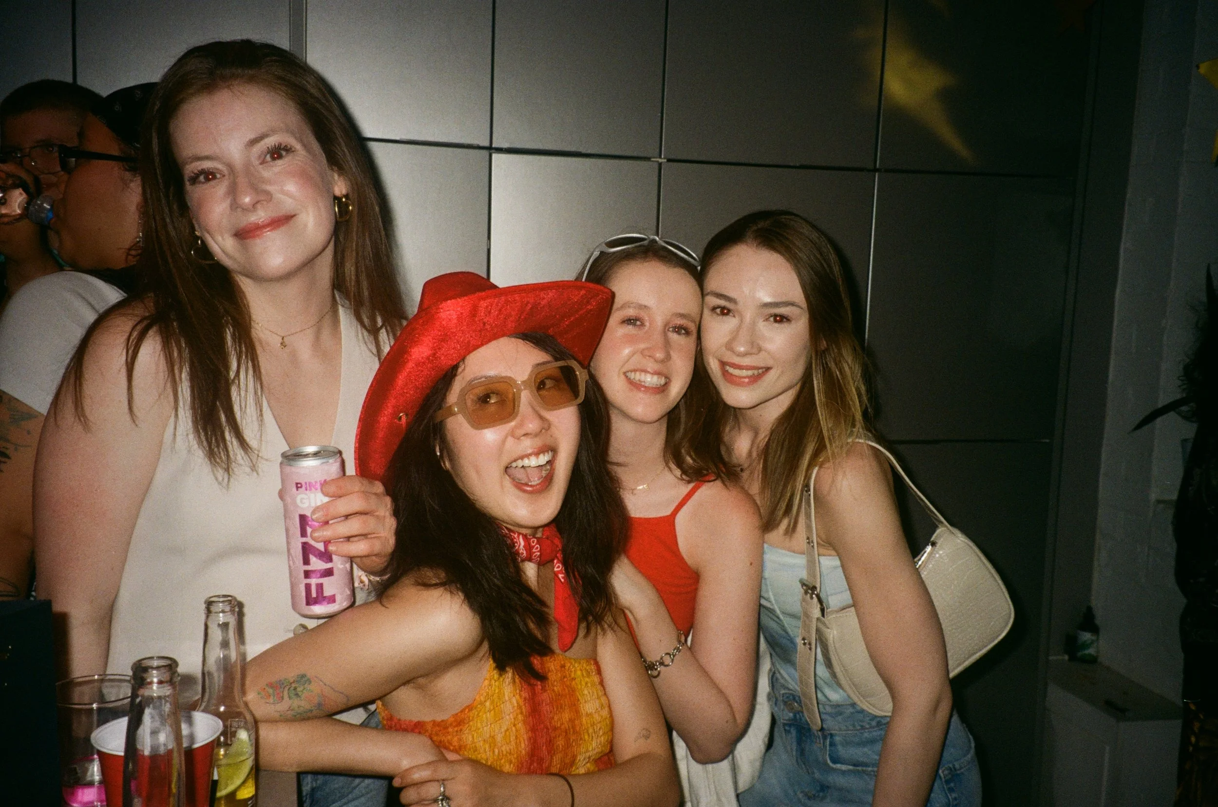 A group of five women celebrating together at a party, smiling and posing for a photo. They are indoors with bottles and drinks on the table.