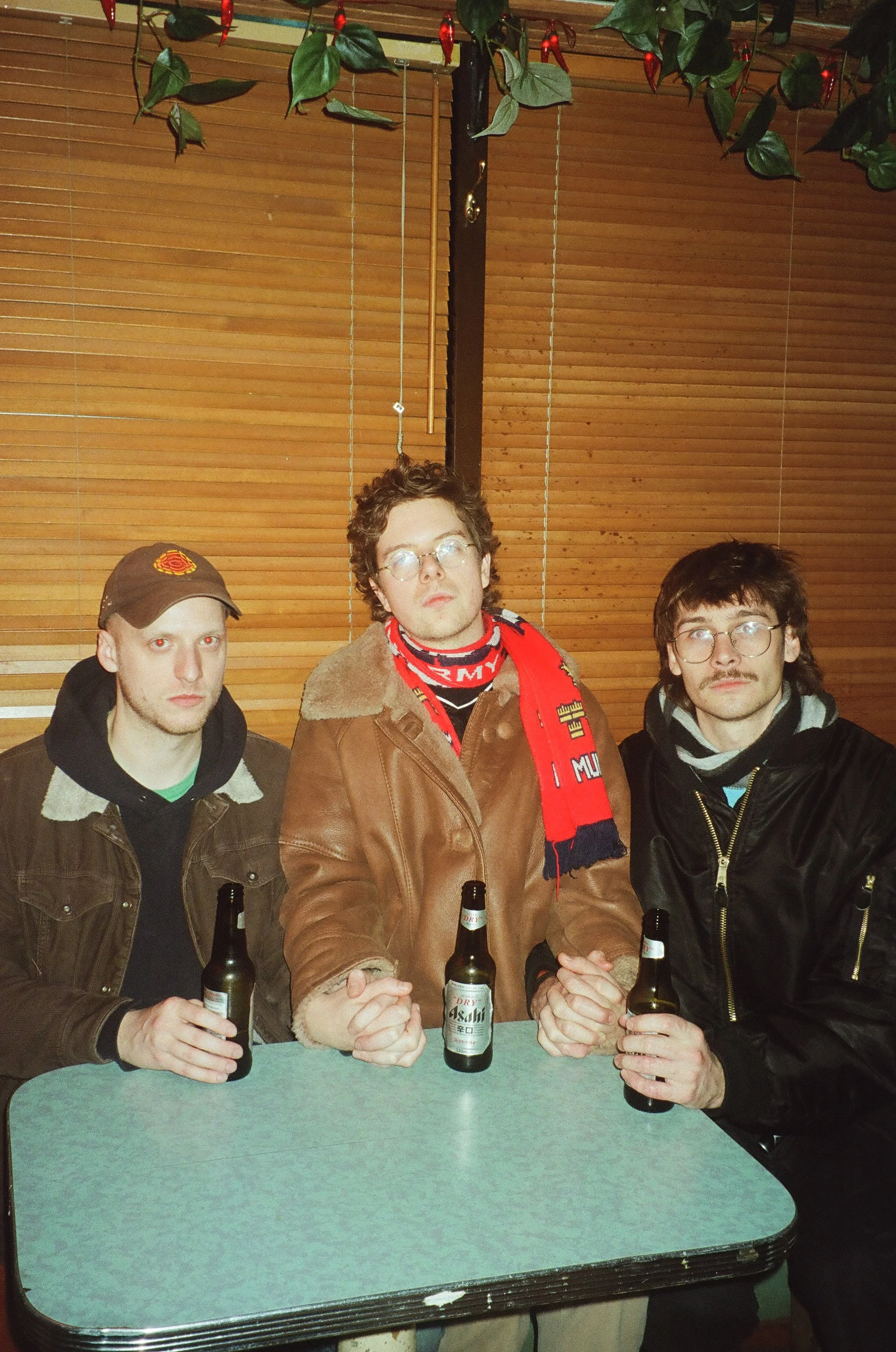 Three young men sitting at a table with beer bottles in front of them in a wooden-paneled room, two of them have glasses and are wearing jackets, one is wearing a baseball cap.