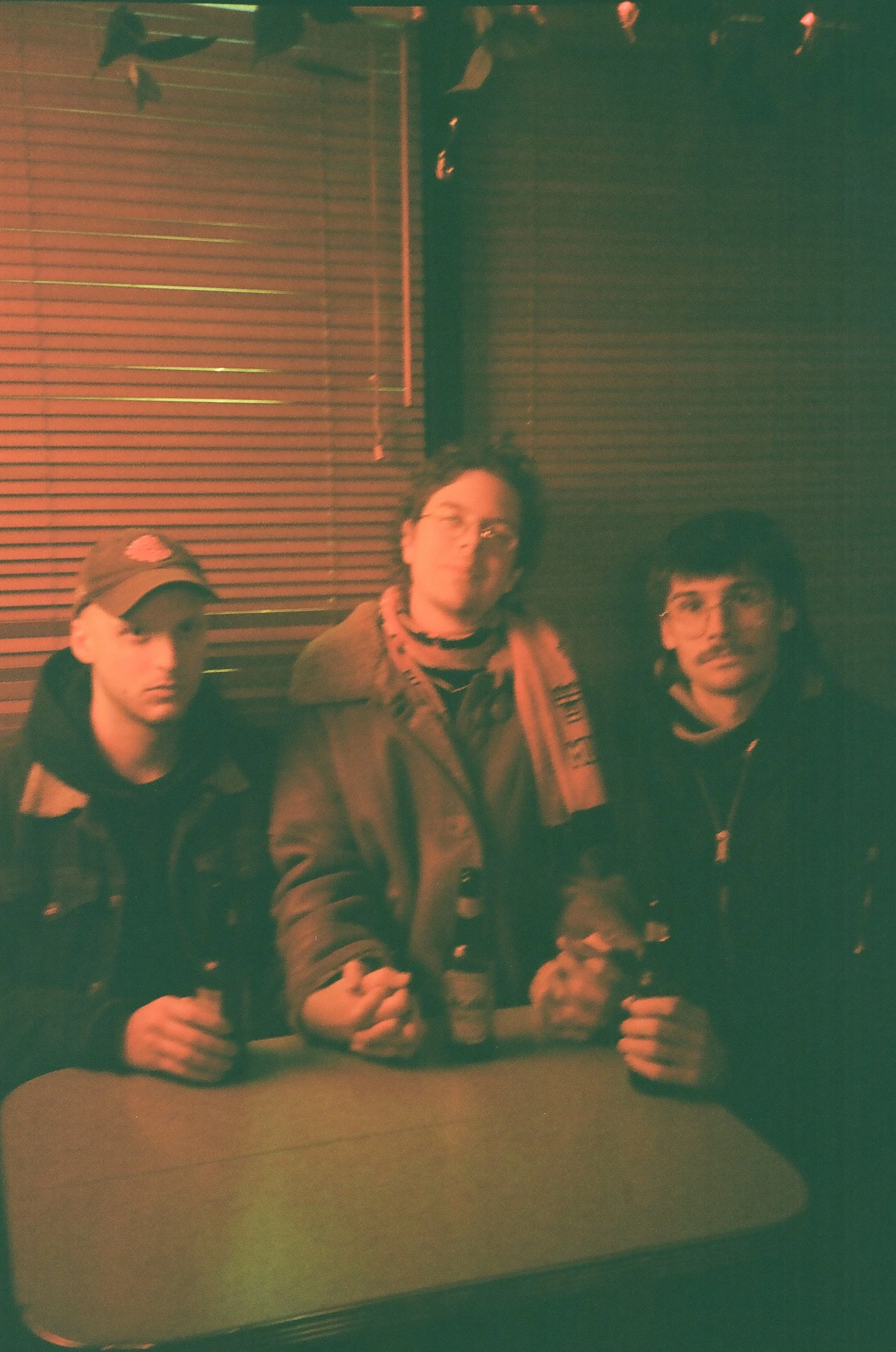 Three young men sitting at a table in a dimly lit room with horizontal striped wall panels. They have drinks in their hands and are looking at the camera.