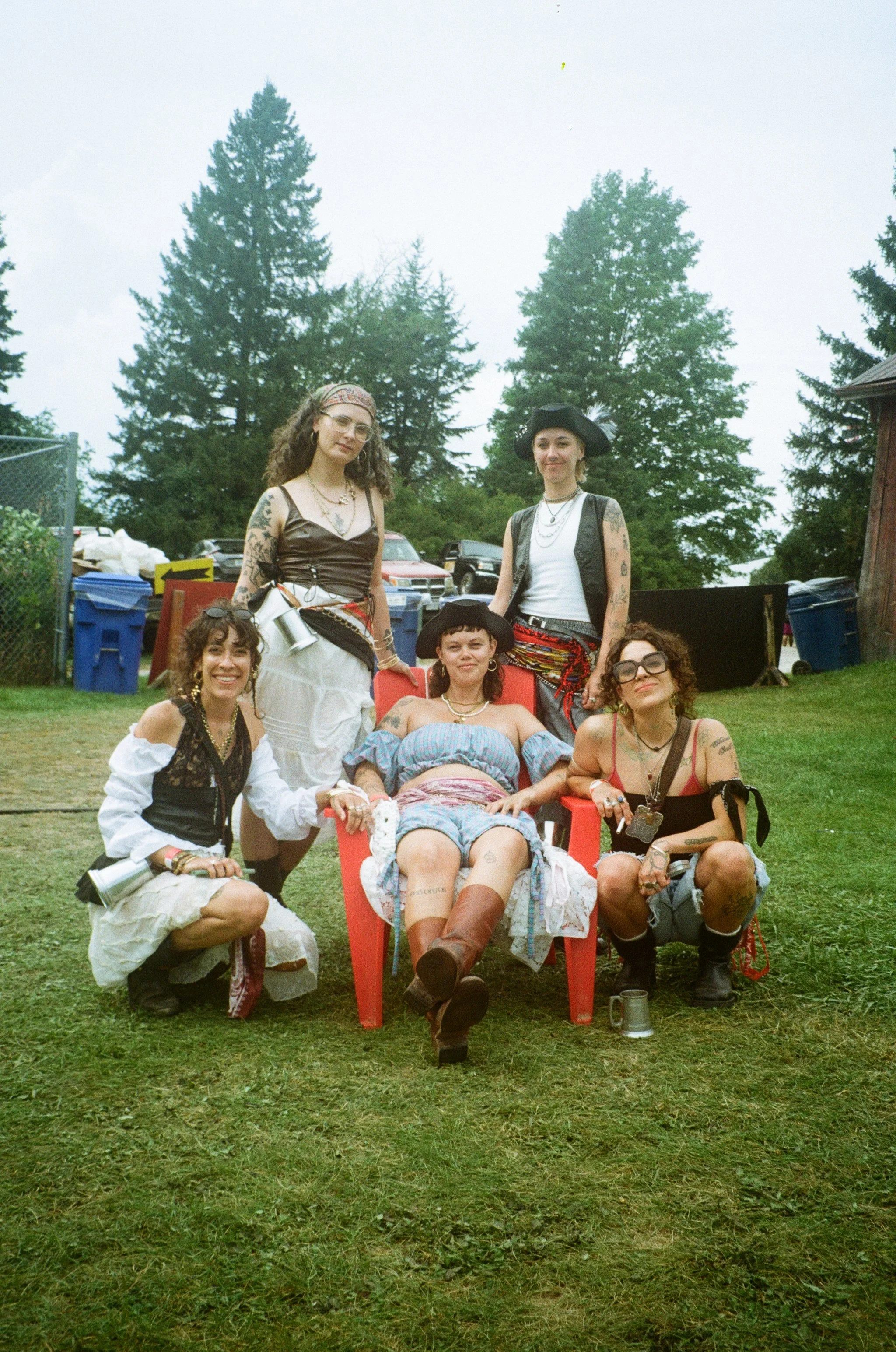 A group of six women with tattoos and wearing bohemian or festival style clothing, posing outdoors on grass with trees in the background, some seated and some standing, with one person in a wheelchair.