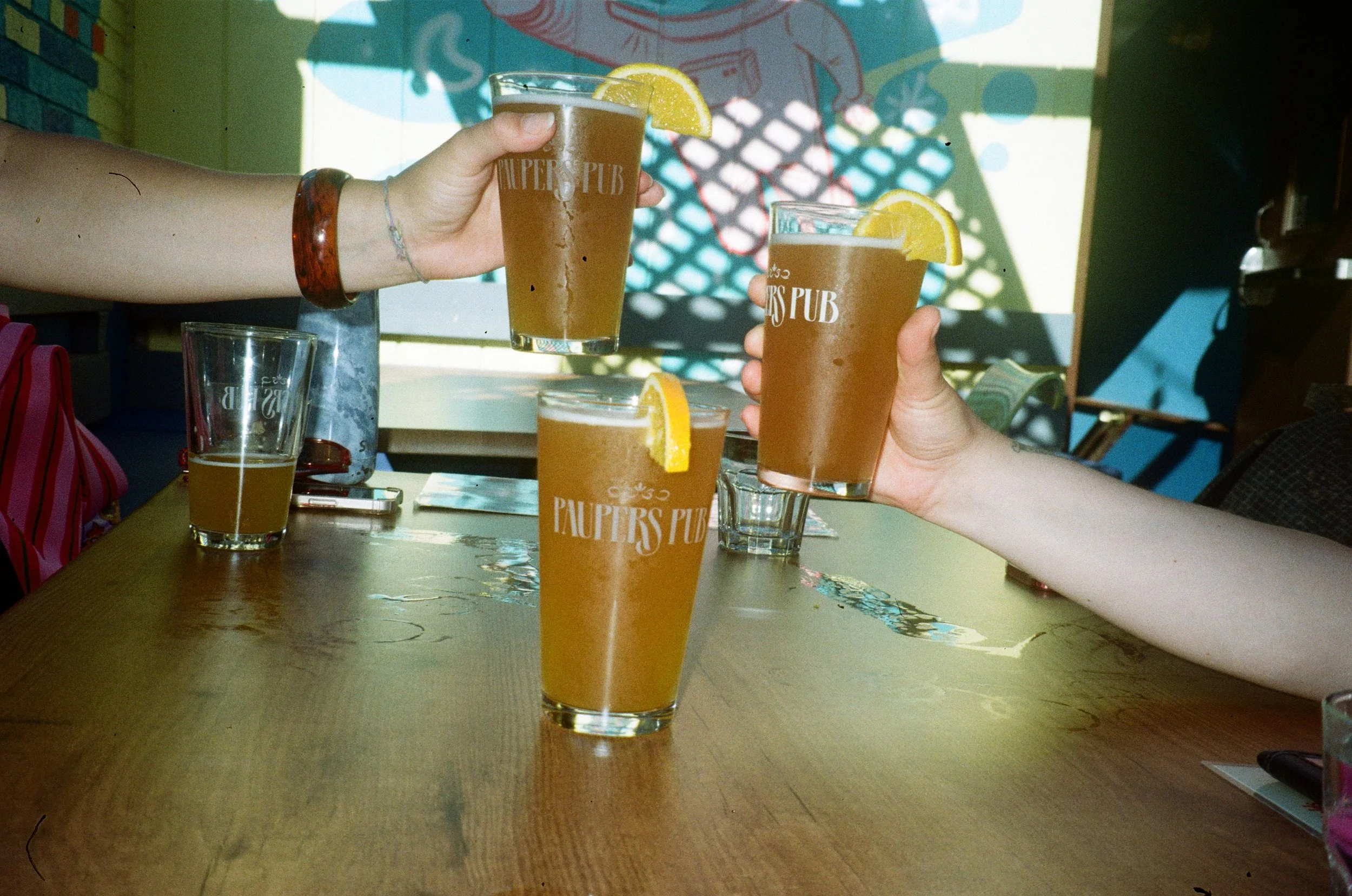Two people are holding beer glasses with lemon wedges, raised in a toast at a table with additional glasses and a colorful background.
