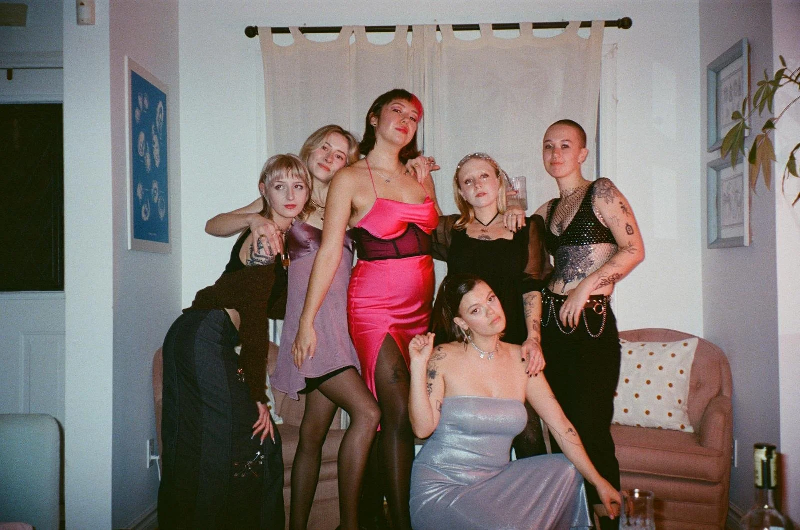 Group of six women in stylish outfits posing in a living room with white walls, artwork, and a pink couch.