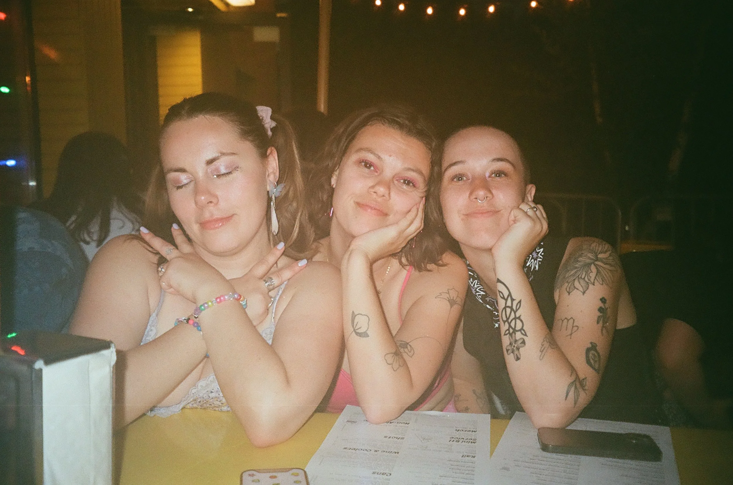 Three women sitting at a table in a dimly lit room, smiling and posing for the photo. They have tattoos on their arms, and are wearing casual clothes and jewelry. There are menus and a cellphone on the table.