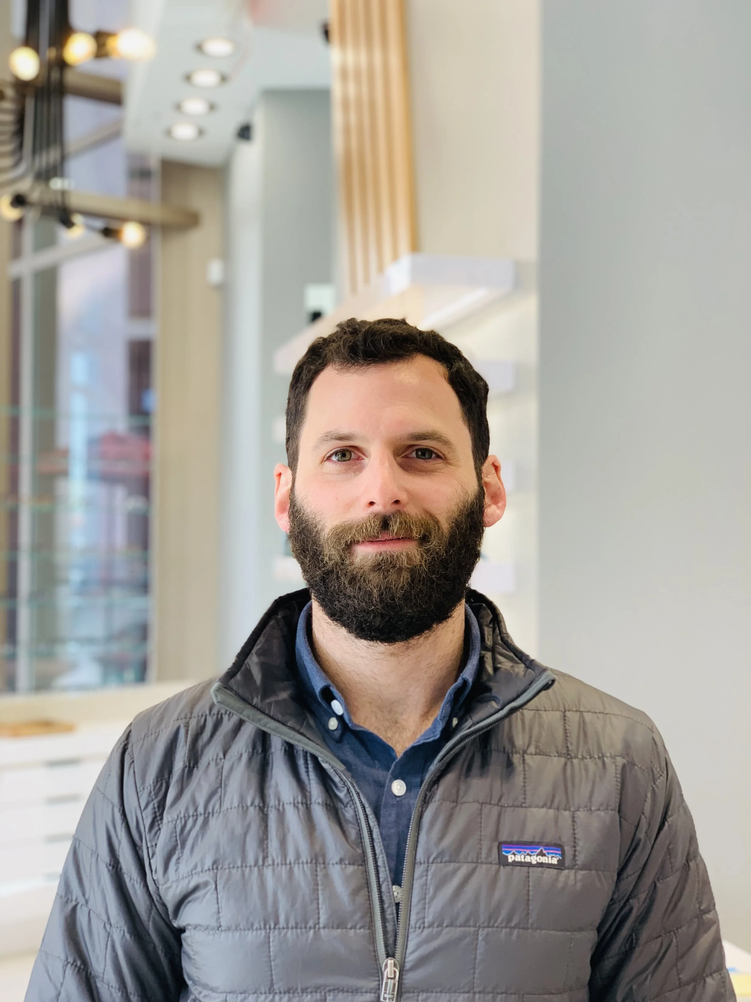 A man with dark hair and a beard wearing a gray Patagonia jacket and a blue collared shirt, standing indoors with a background of modern interior design including wooden panels, lights, and glass windows.