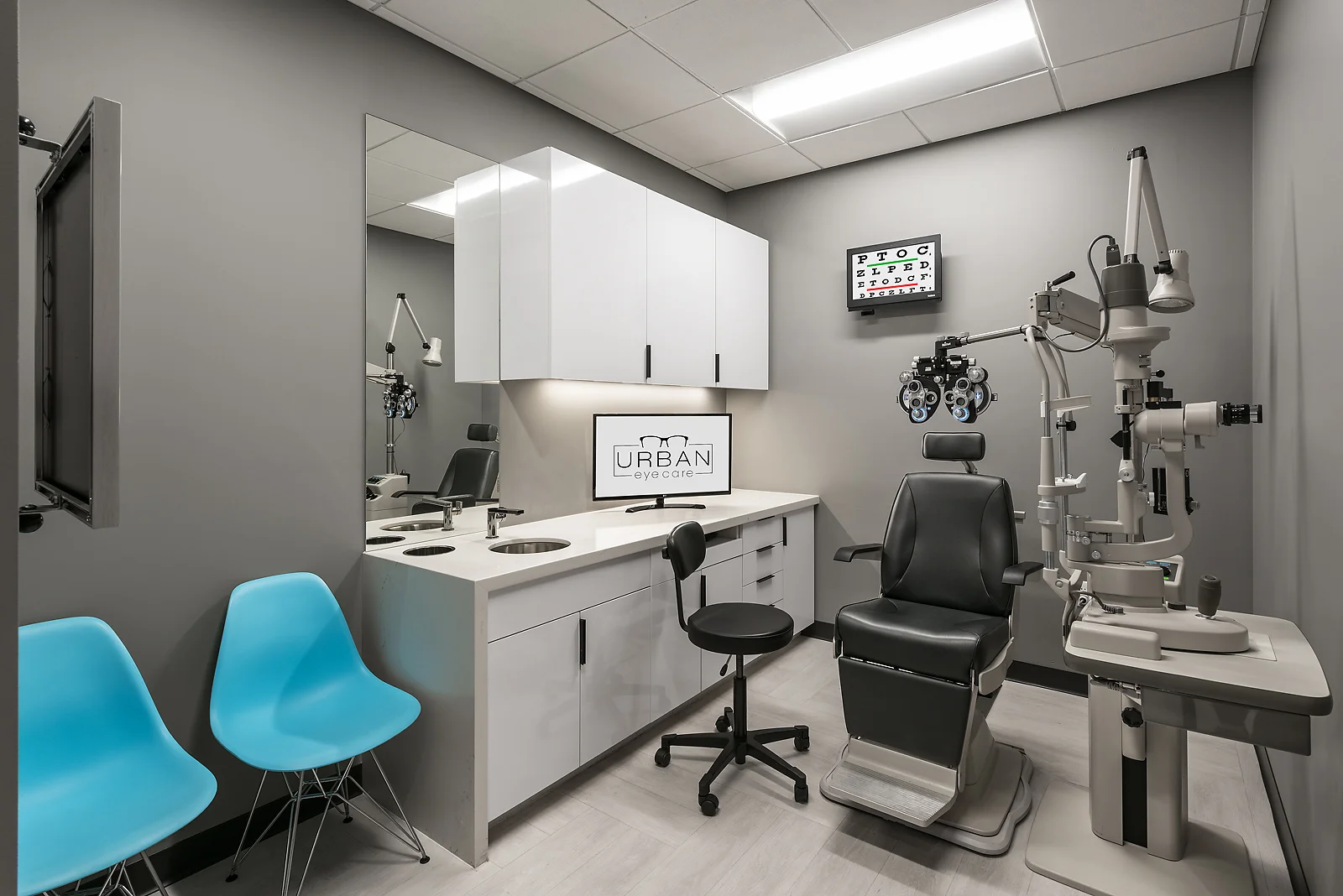 A modern eye care exam room with gray walls, white cabinets, and medical equipment, including an eye examination machine, a chair for the patient, and a display screen with the 'URBAN eye care' logo.