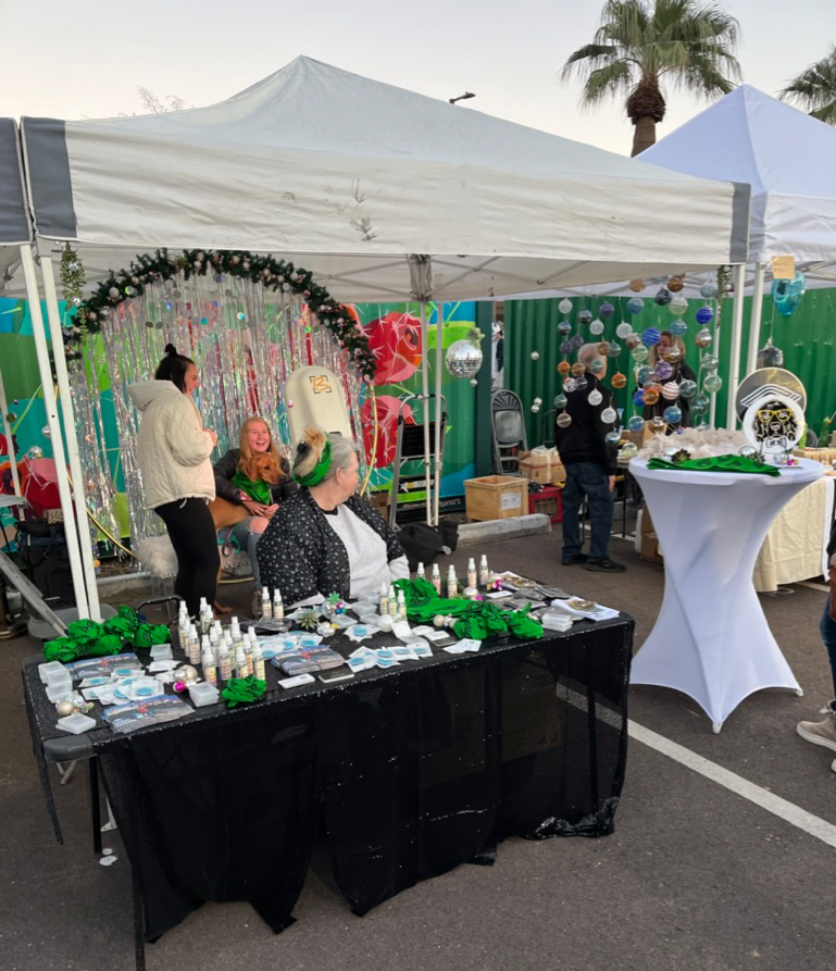 Outdoor market stall with various items including small bottles, green fabric, and decorations. Two women are seated behind the table, one with green hair, and a woman stands nearby. Other tents and palm trees are visible in the background.