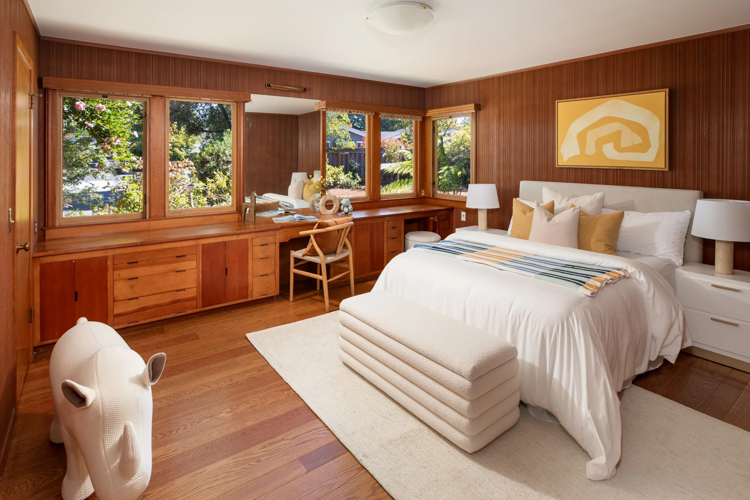 A cozy bedroom with wood-paneled walls and large windows overlooking a lush garden. The bed has white bedding and multiple throw pillows, with a beige upholstered bench at the foot. Nearby nightstands hold white lamps, and a wooden desk with a chair 