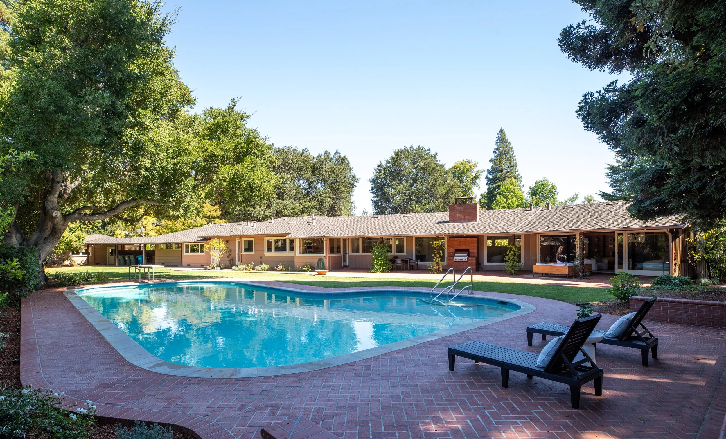 A backyard with a swimming pool, surrounded by red brick paving and lounge chairs. Behind the pool, there is a single-story house with a patio, a chimney, and large windows. Tall trees with lush green foliage shade the yard, and the sky is clear and 