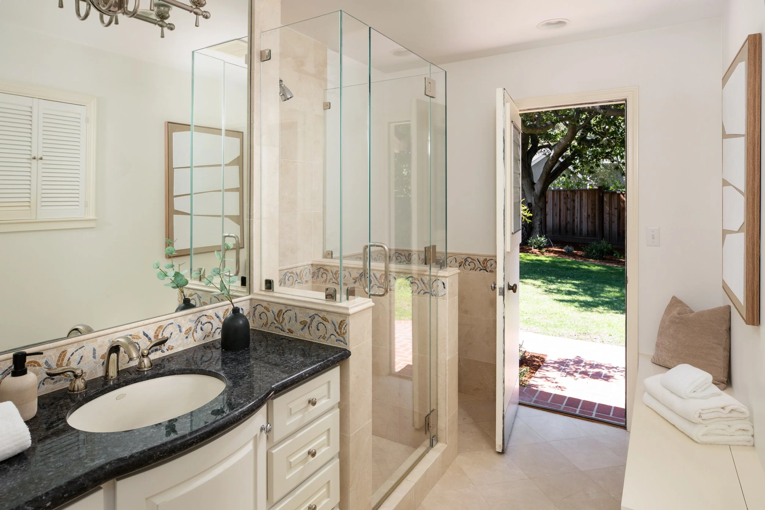 A bathroom with a black granite countertop sink, white cabinets, a mirror, and a glass shower enclosure. The bathroom opens to a backyard with a tree and a fenced yard.