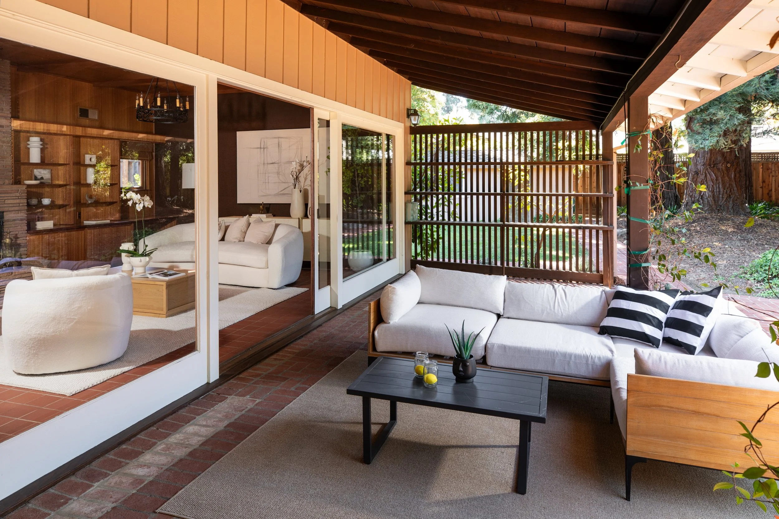 A cozy living room with beige and white furniture, including a round chair and sofa, decorated with black and white striped pillows. A black coffee table has small plants and candles. The room has large glass sliding doors opening to a backyard with 