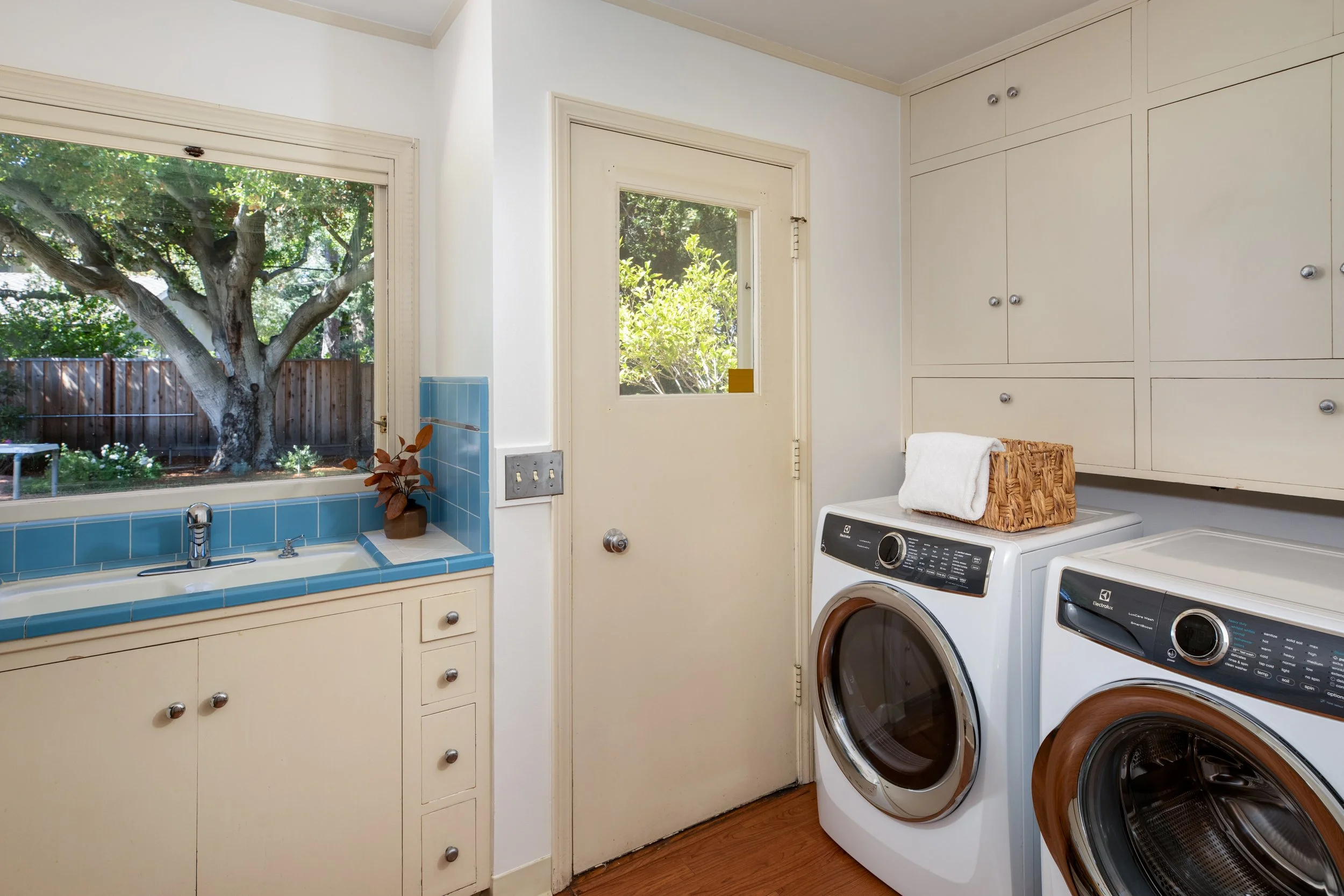 Laundry room with two front-loading washing machines, beige cabinets, a window showing a large outdoor tree, and a basket of folded towels on top of one of the machines.