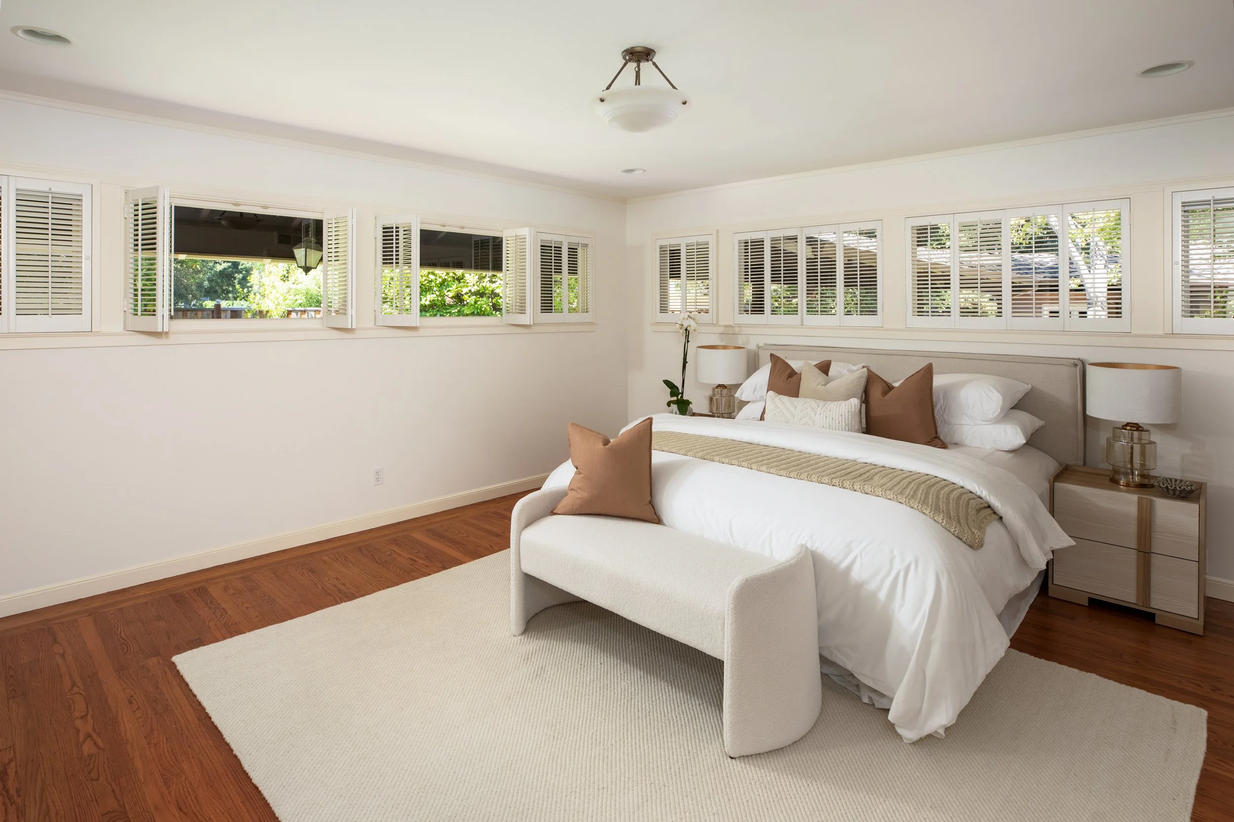 A bedroom with white walls, hardwood flooring, and multiple windows with white shutters. The bed has white bedding, brown and cream pillows, and a beige throw blanket. There are two bedside tables with lamps, and a small potted plant is near the wind