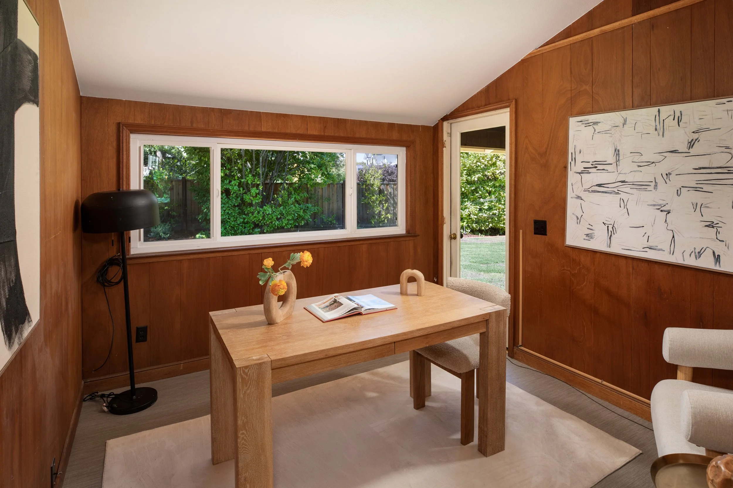 A cozy dining room with wood-paneled walls, a rectangular wooden dining table with chairs, a large window showing green trees outside, a black floor lamp, and abstract artwork on the wall.