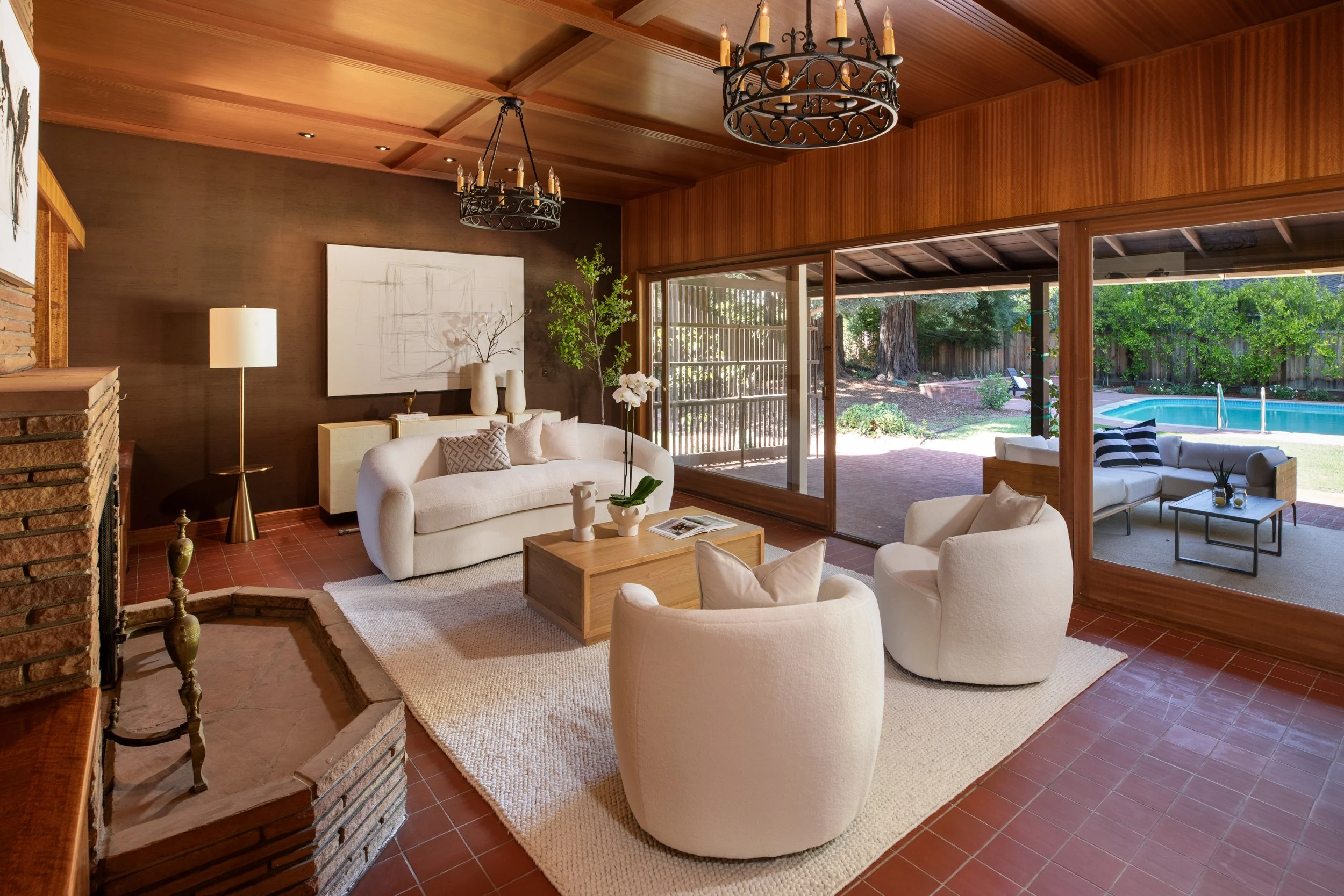Living room with white sofas, a coffee table, a floor lamp, and wall art. Large glass doors lead outside to a patio with a pool and greenery.