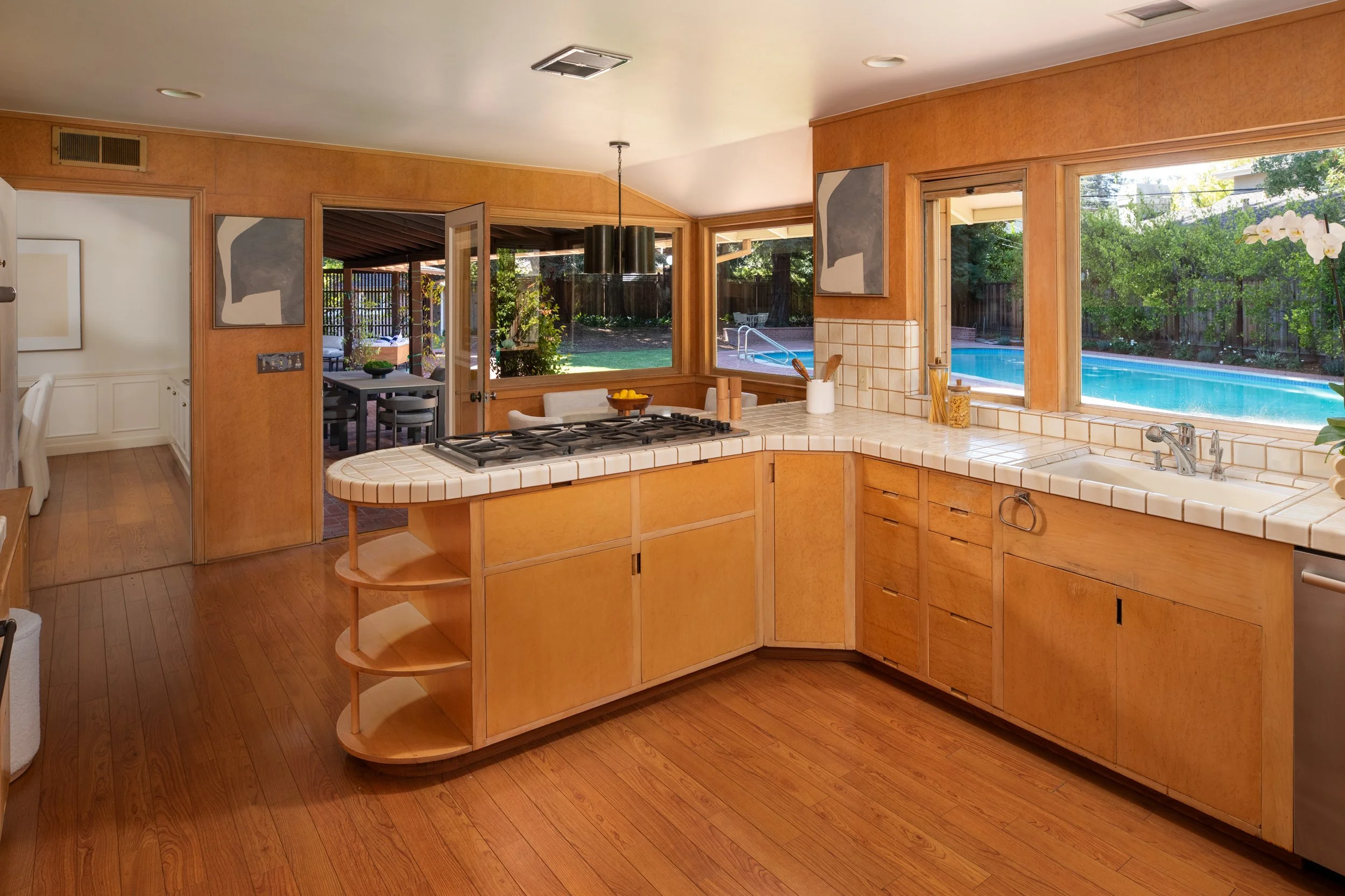Kitchen with wooden cabinets and tile countertop, large windows showing backyard with swimming pool and lush greenery, dining area outside, modern art on walls, and hardwood flooring.