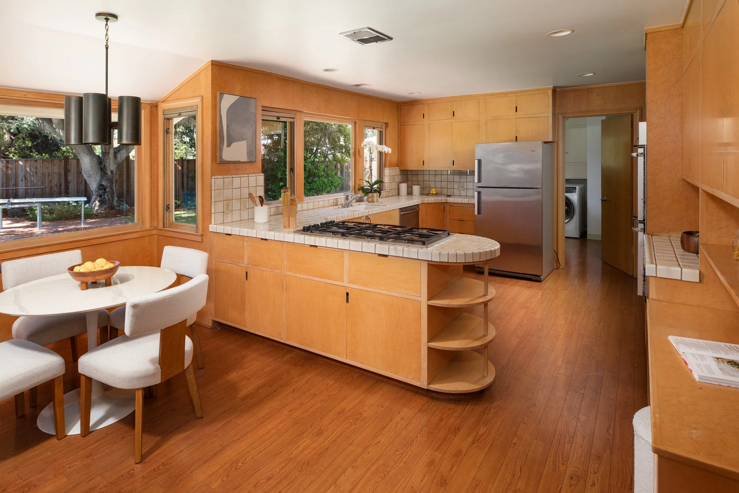 View of a cozy kitchen with wooden cabinets, large windows looking outside, a white round dining table with beige upholstered chairs, and modern ceiling lighting fixtures.