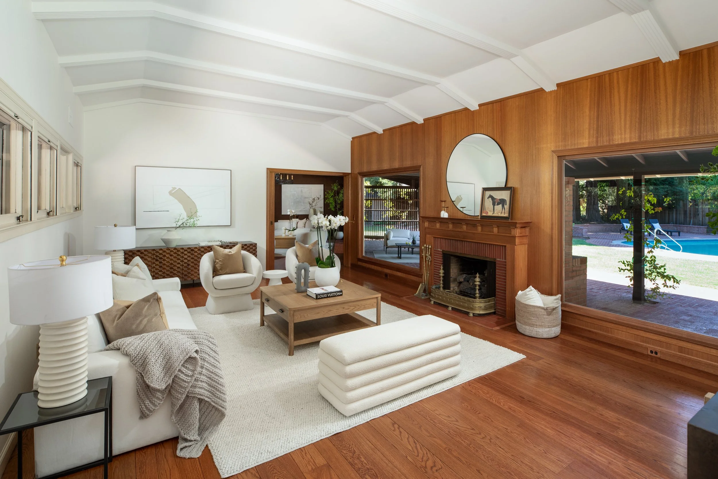 Living room with white sofa, beige pillows, wood coffee table, lamps, and artwork, with view of backyard pool through large windows.