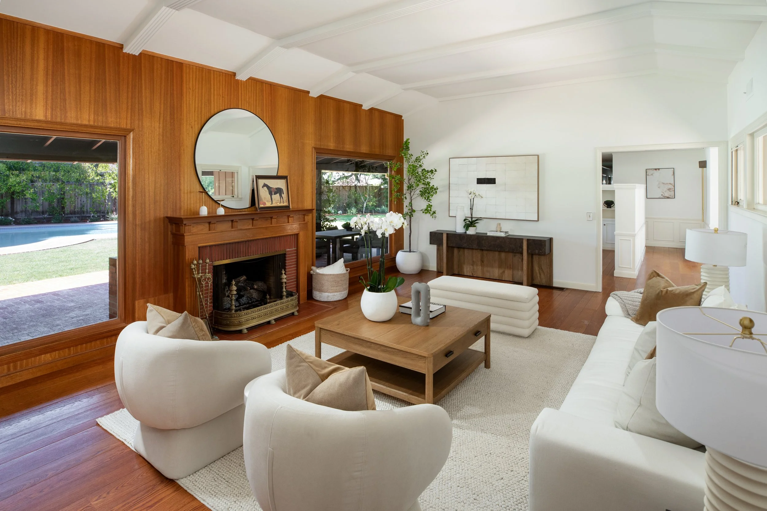 Living room with white sofas, wooden coffee table, wood-paneled wall, large mirror above fireplace, potted plant, artwork, and sliding glass door revealing backyard pool and greenery.