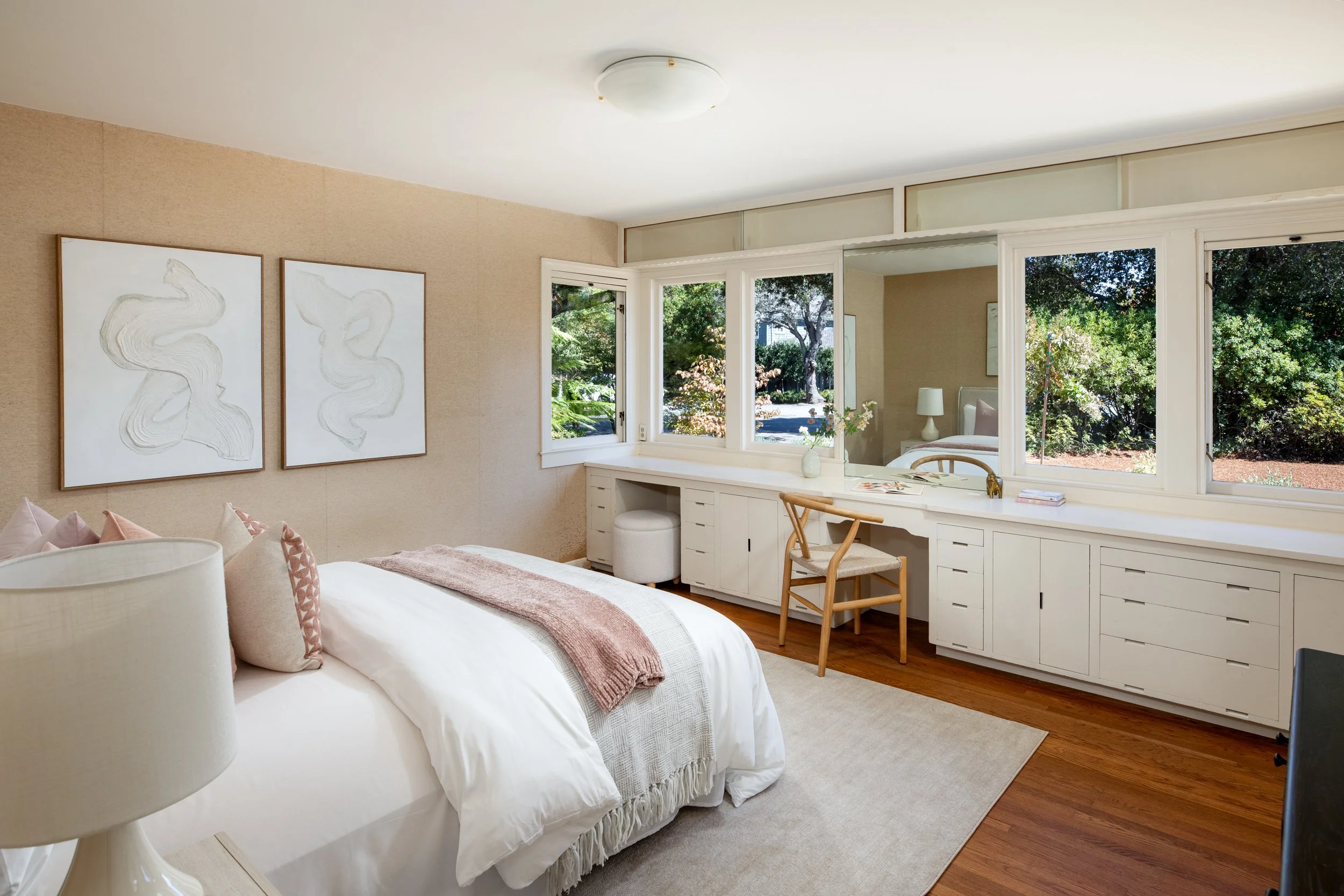 Bright bedroom with a bed, pink and white pillows, bedside lamp, artwork on beige wall, window with a view of trees, white desk with a vase and a chair.