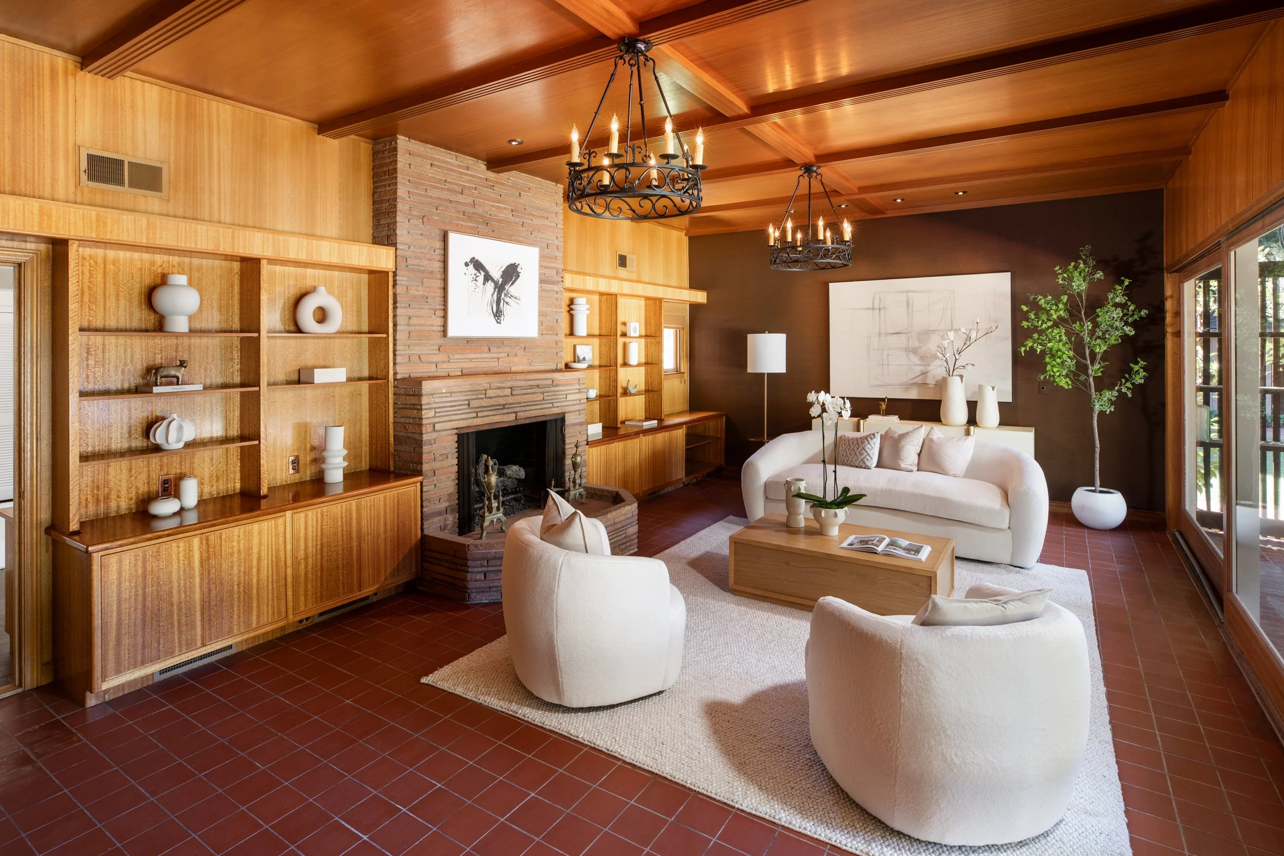 Living room with wooden ceiling and walls, brick fireplace, beige sofas, white vases, artwork, large windows, and chandeliers.