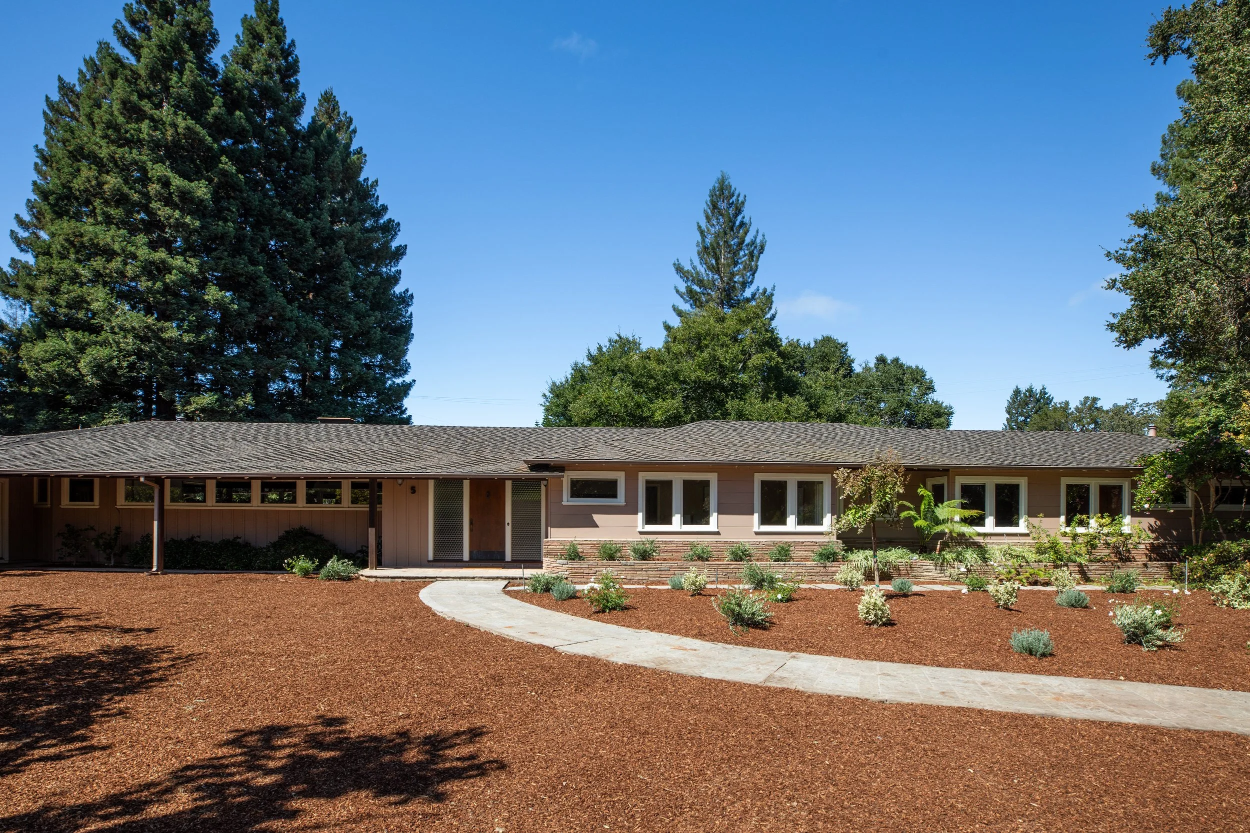 A single-story house with a light-colored exterior and dark roof shingles, surrounded by a well-maintained yard with a curved concrete pathway, small plants, and mulch, under a clear blue sky with tall green trees in the background.