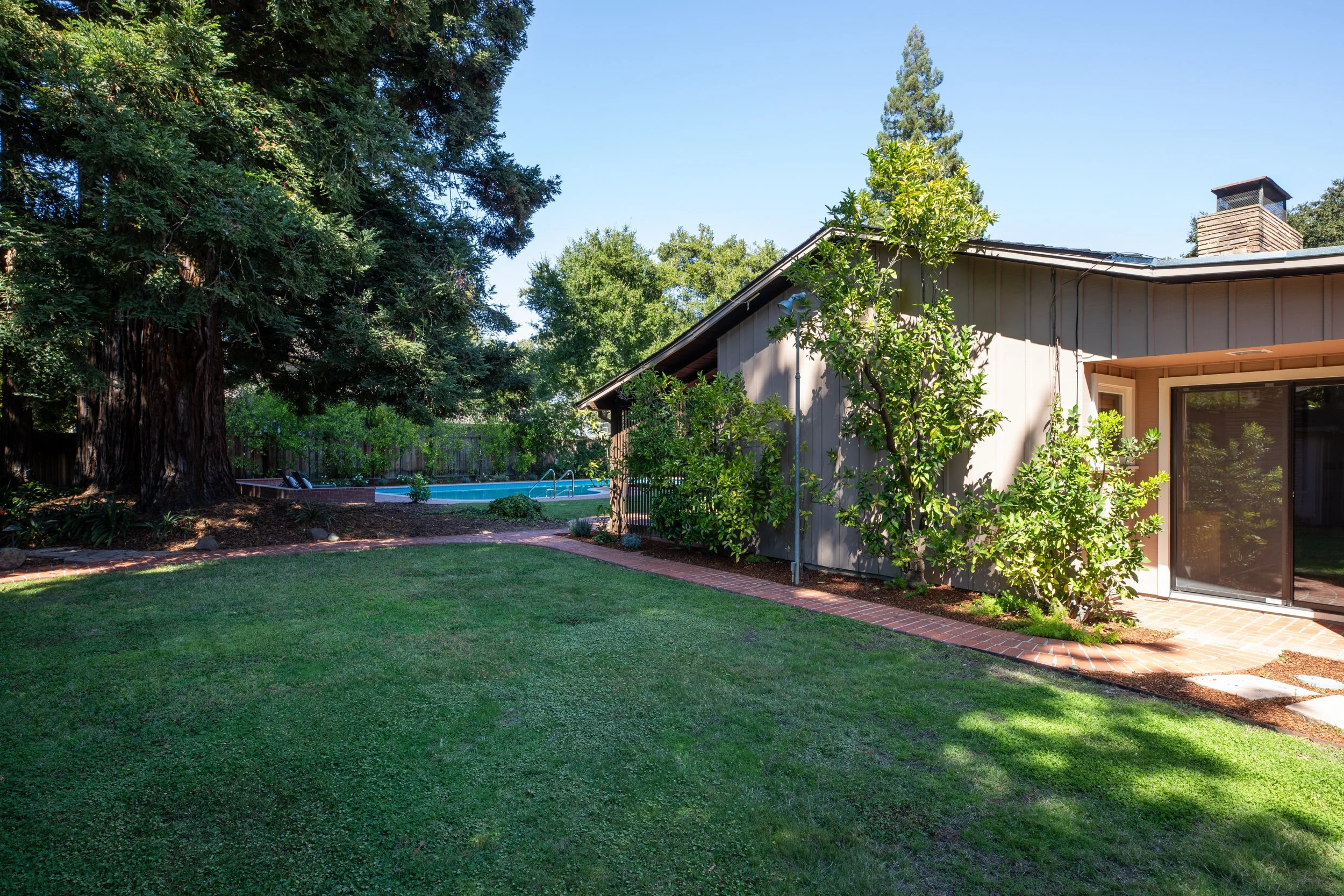 A backyard with a green lawn, trees, a swimming pool, a brick pathway, and a house with a gray exterior and large windows.