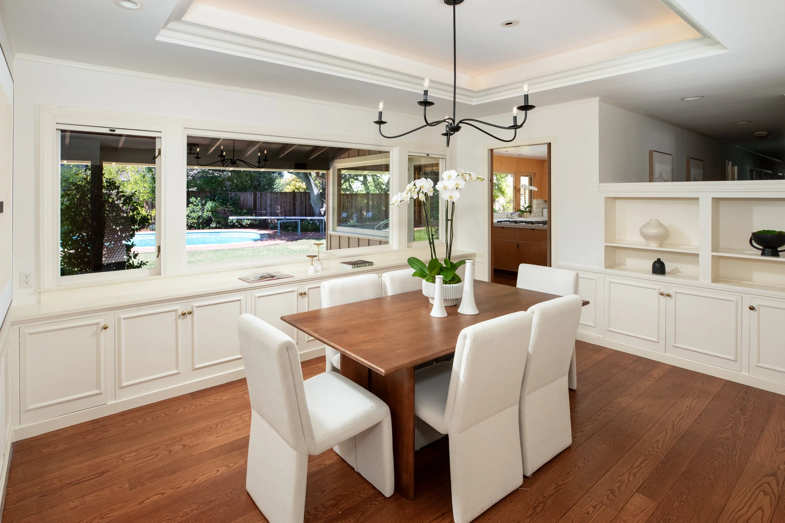 A dining room with a wooden table surrounded by six cream-colored upholstered chairs, a white orchid plant on the table, large windows showing a backyard with a pool, and a hutch with decorative vases.