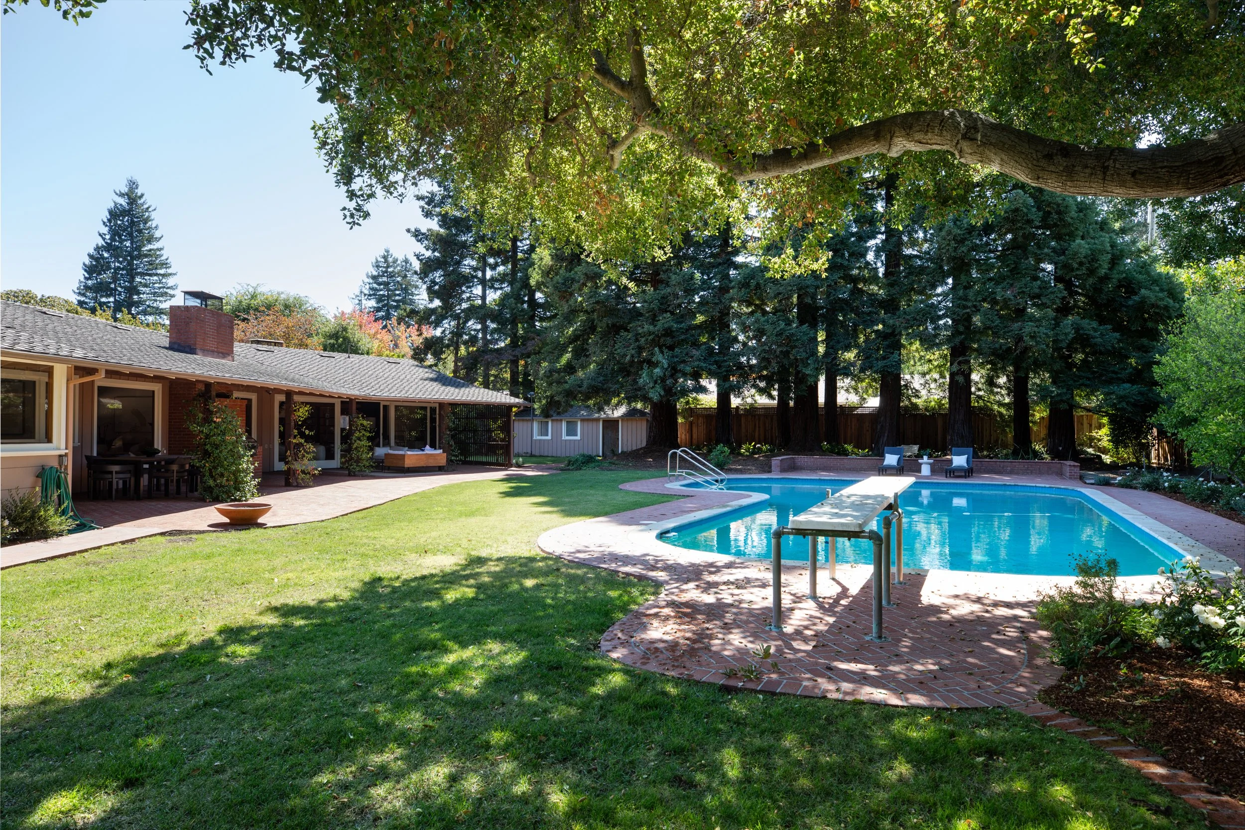 Backyard with a swimming pool, patio with outdoor furniture, trees, and a house with a brick chimney under a clear blue sky.